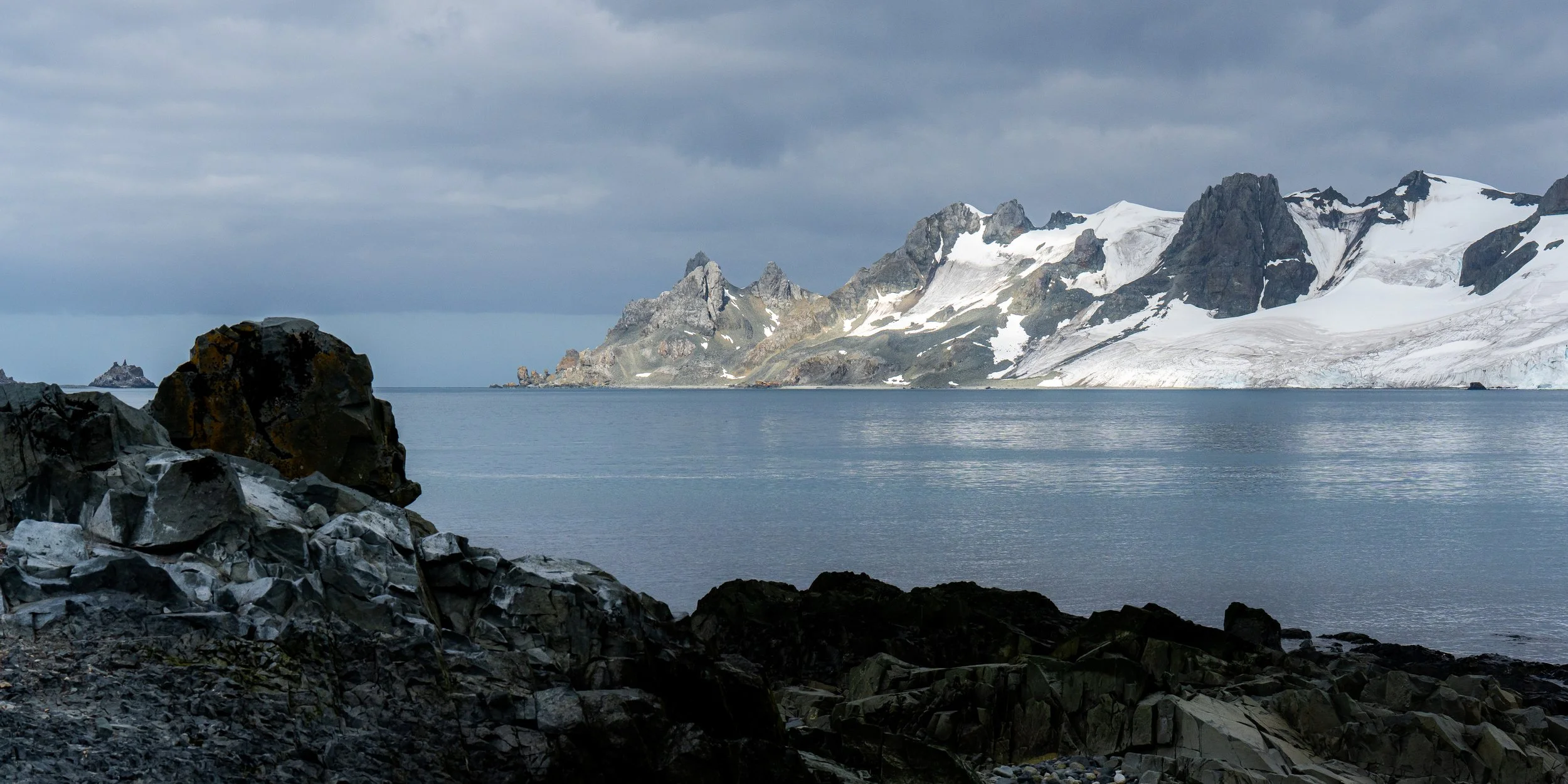 Mountains with snow and glaciers beside a body of water, with rocks in the foreground and a cloudy sky.