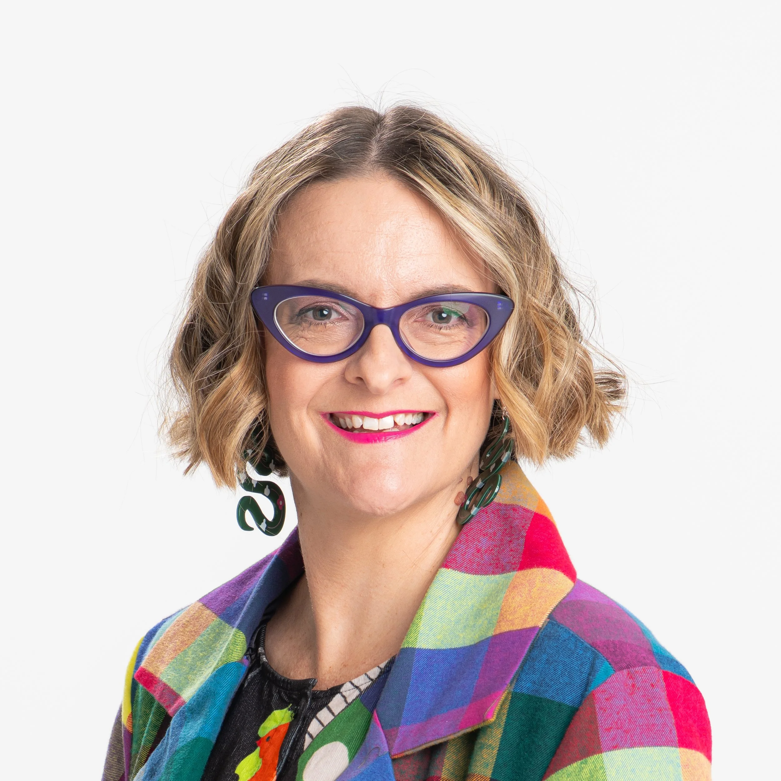 Close-up of a woman with light brown wavy hair wearing purple glasses and a rainbow-colored checkered jacket, smiling against a white background.
