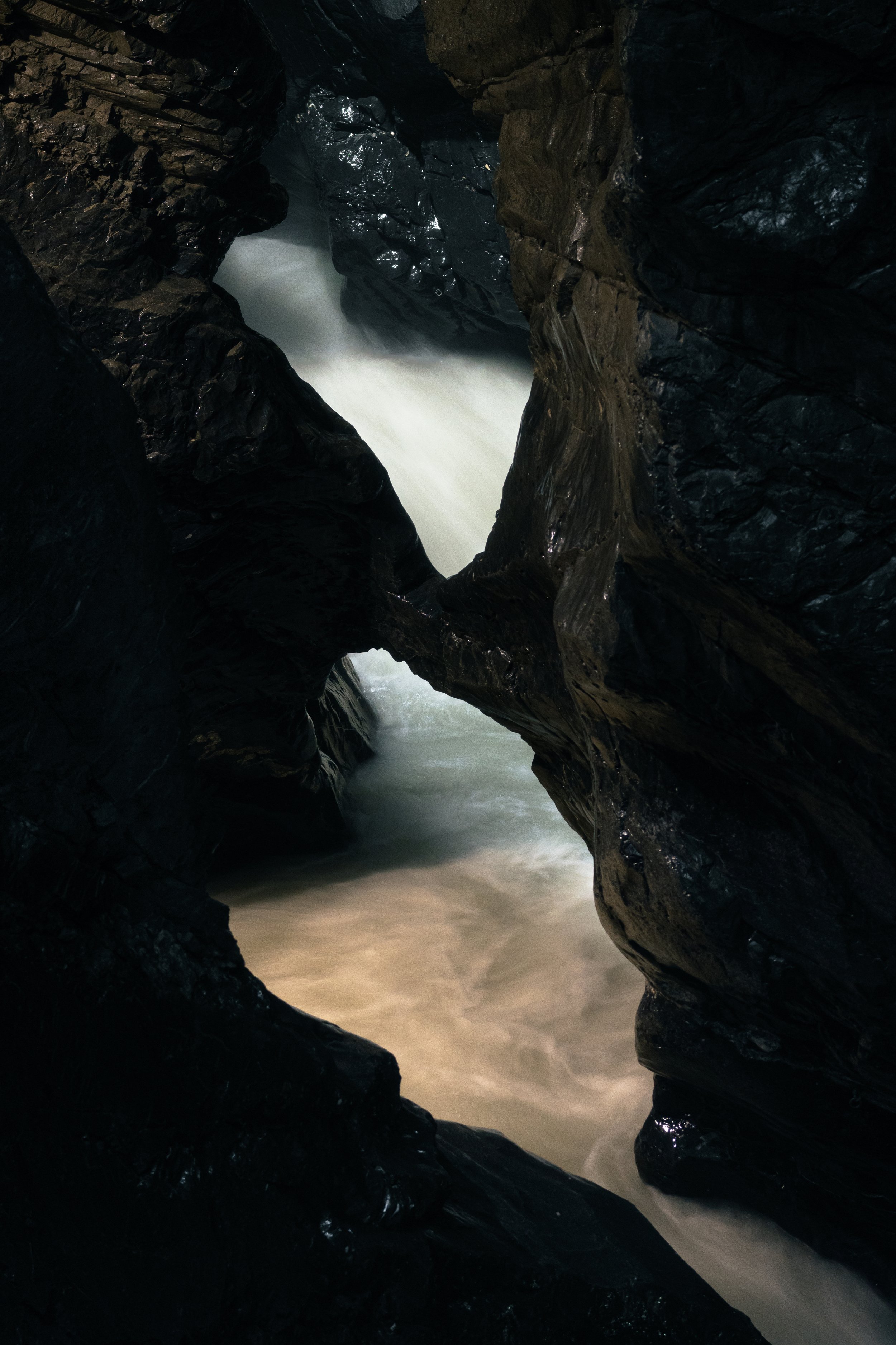 Water rushing through a narrow, dark rocky canyon with glossy black rock walls and white water.
