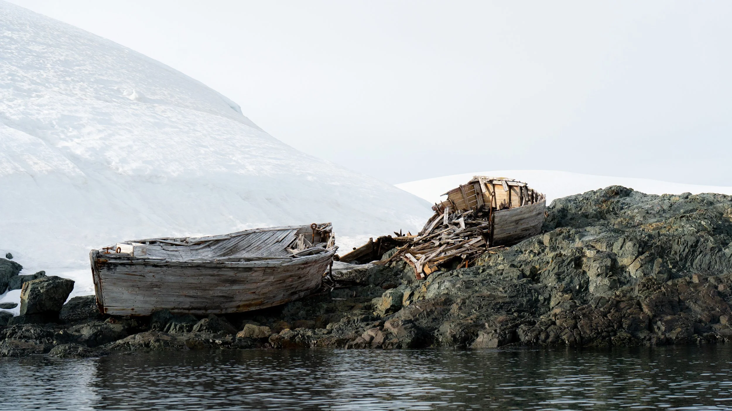 Two abandoned wooden boats on rocky shore with snow-covered mountains in the background.