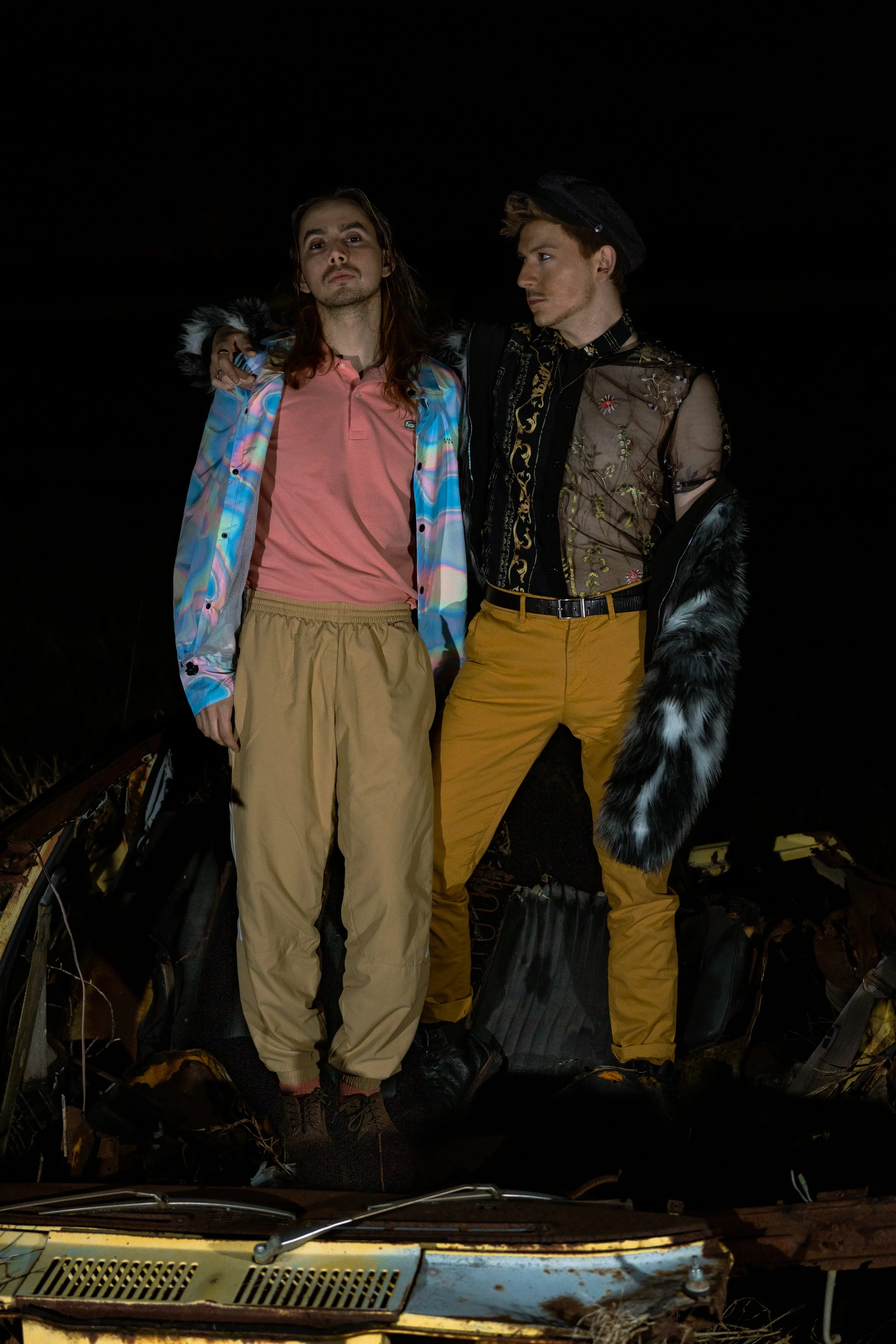 Two young men standing close together at night, one with long hair wearing a pastel-colored jacket and beige pants, the other with short hair wearing an embroidered sheer top, tan pants, and a black beret, with black and white furry coat draped over 