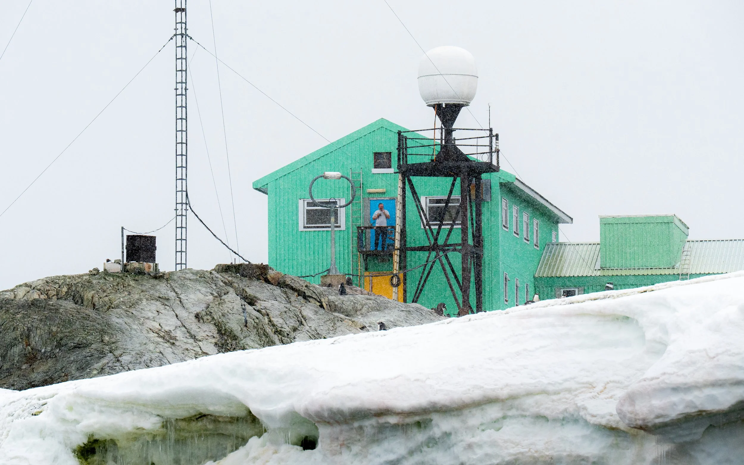 A person standing on a balcony of a green building with antennas and satellite dishes, snow-covered ground, and a rocky foreground, during snowy weather.