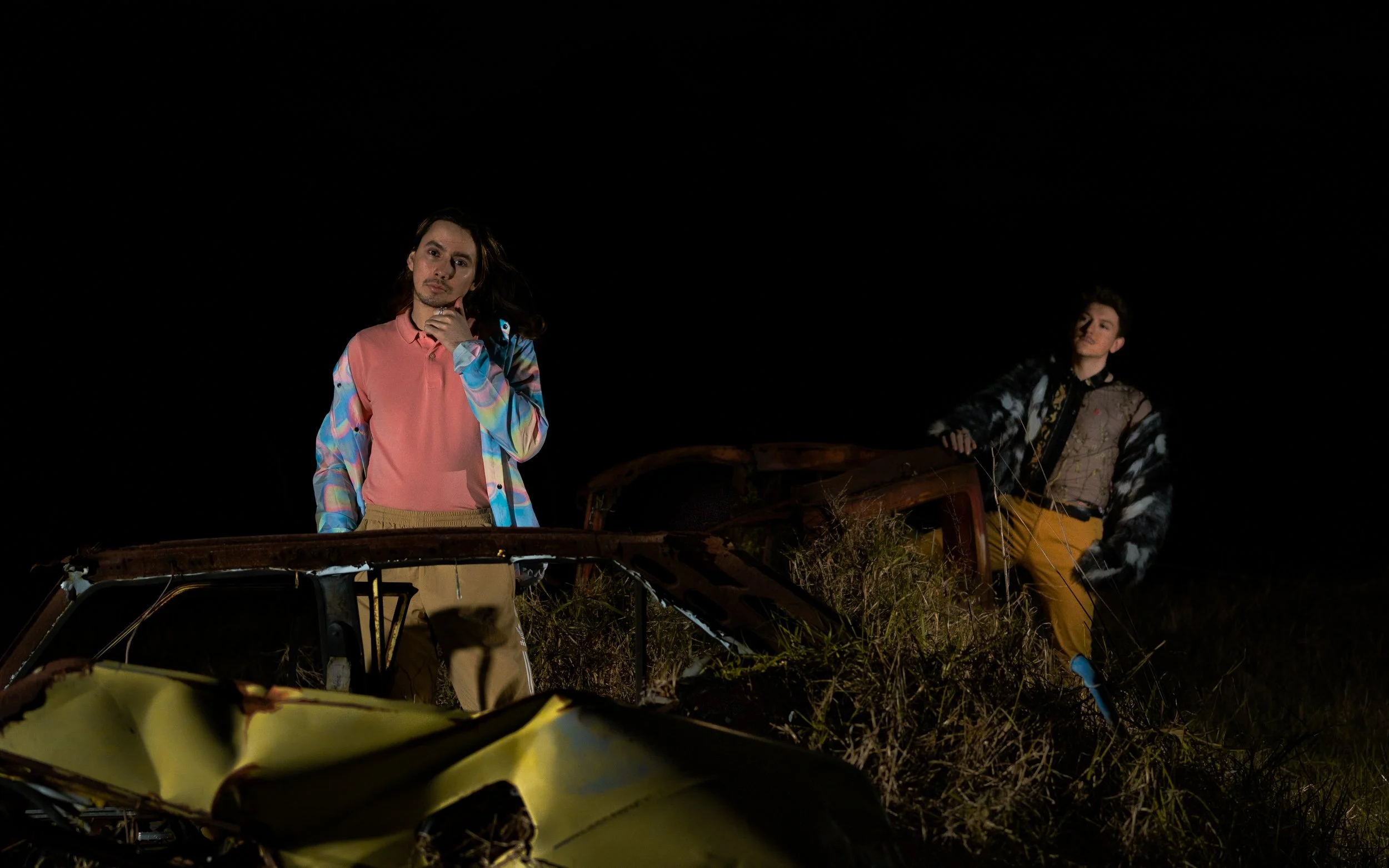 Two young men standing in front of an abandoned, rusty car at night. One has long hair and is wearing a colorful, patterned jacket, and the other has short hair and is wearing a patterned jacket. Both are looking at the camera.