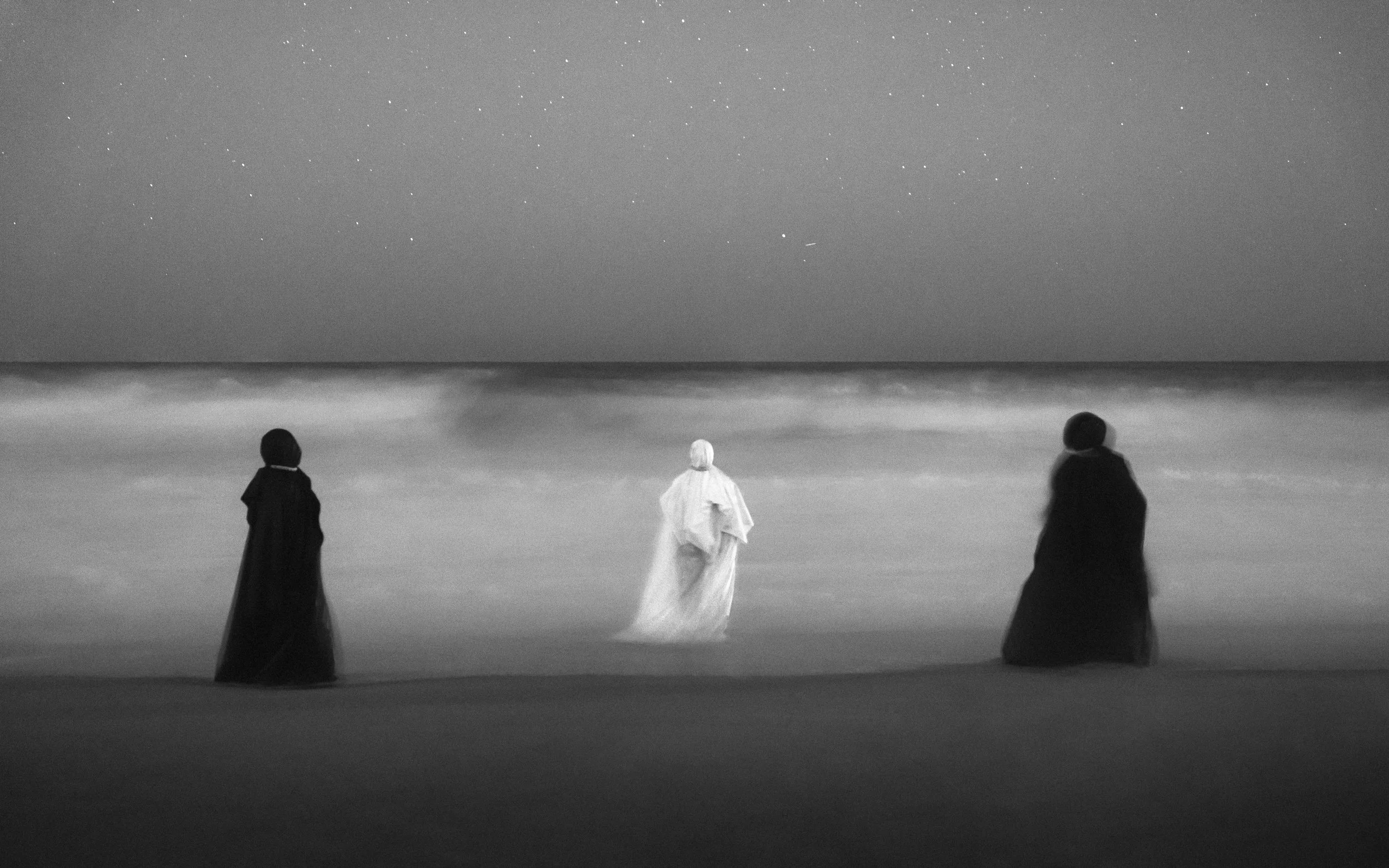 Three people in dark cloaks standing on a beach at night, facing the ocean with stars visible in the sky.
