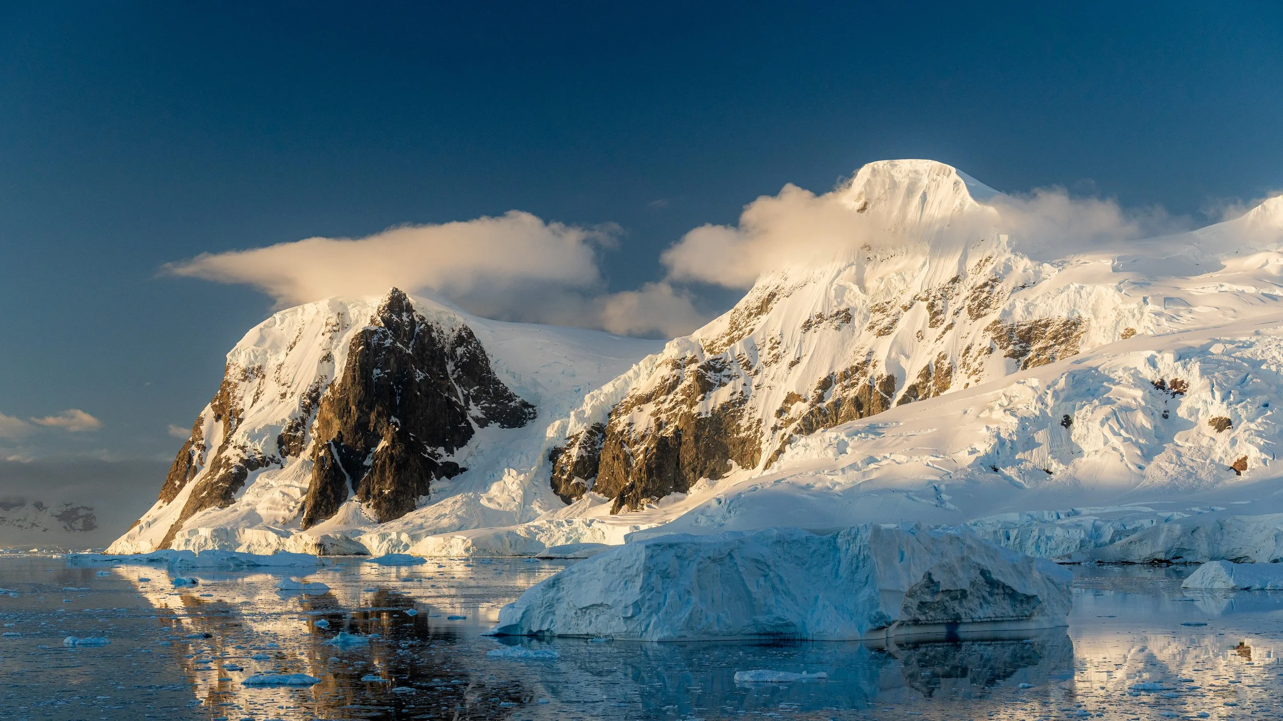 Snow-covered mountain peaks with glaciers, partly obscured by clouds, reflected in calm icy water below.