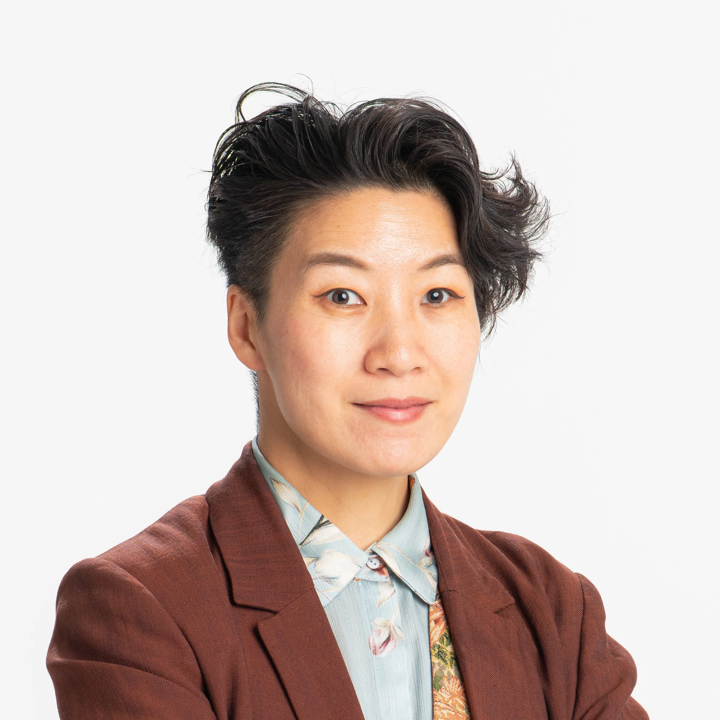A woman with short dark hair wearing a brown blazer, a patterned shirt, and a light blue shirt underneath, smiling softly against a plain white background.