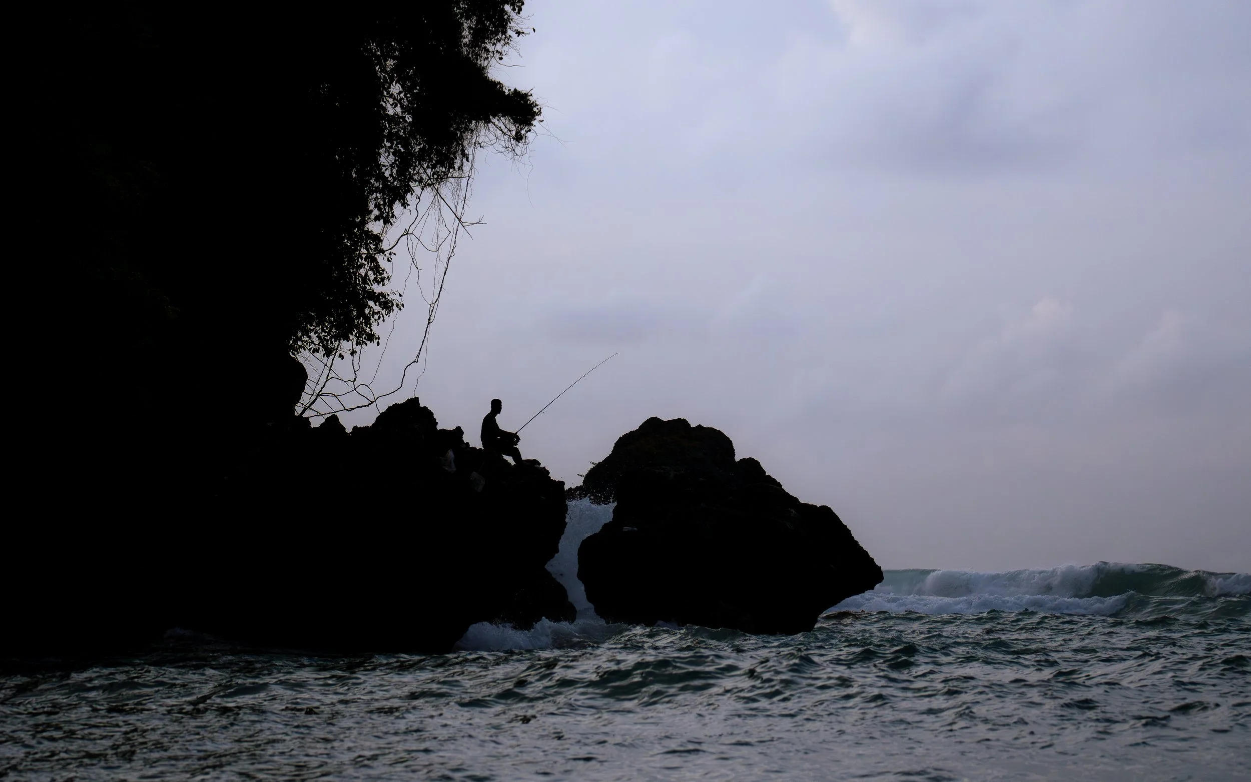 Silhouette of a person fishing on the rocky coast during overcast weather with ocean waves crashing below.