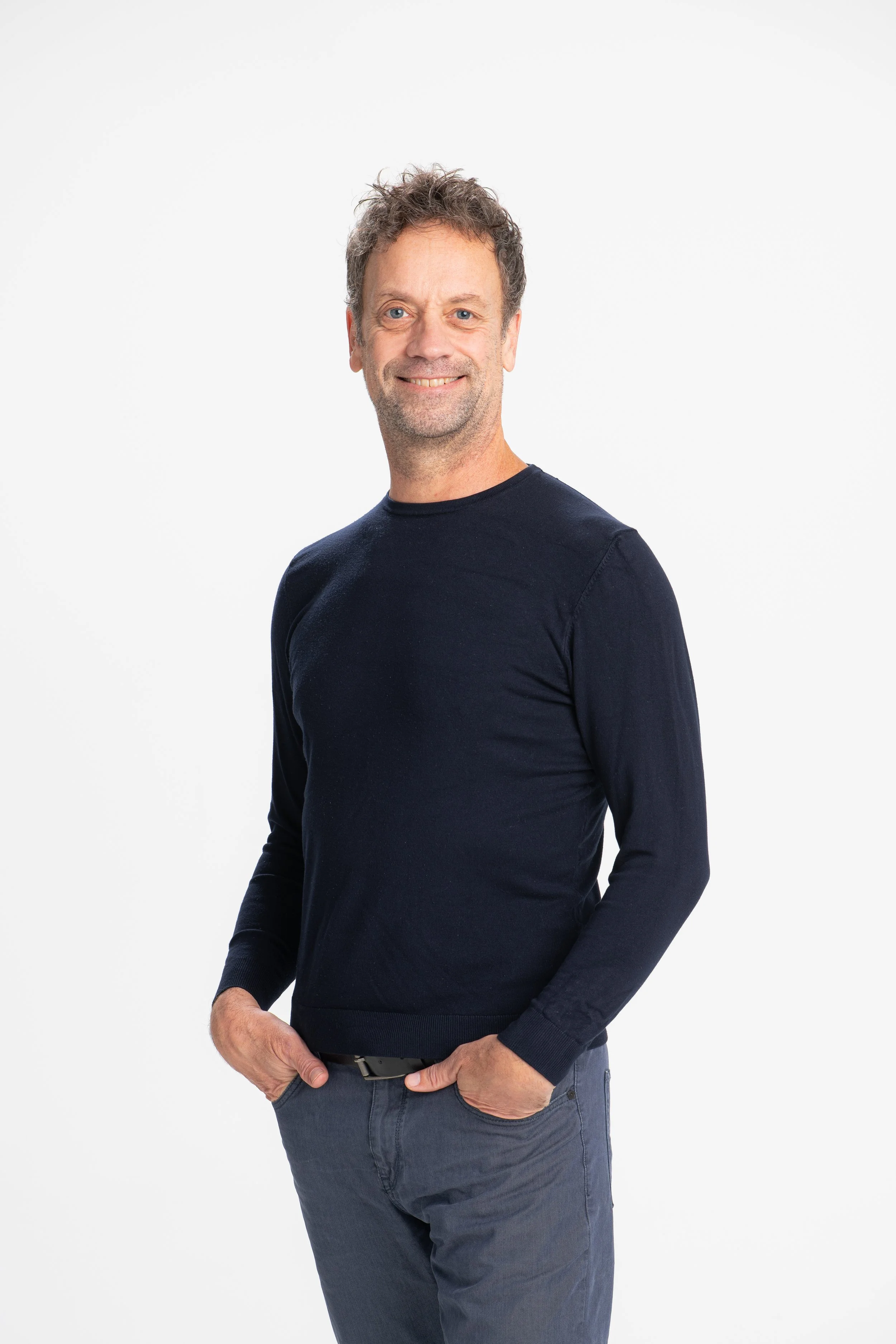 A middle-aged man with curly brown hair and blue eyes is smiling and looking at the camera. He is wearing a navy blue long-sleeve shirt and gray jeans, standing against a plain white background.