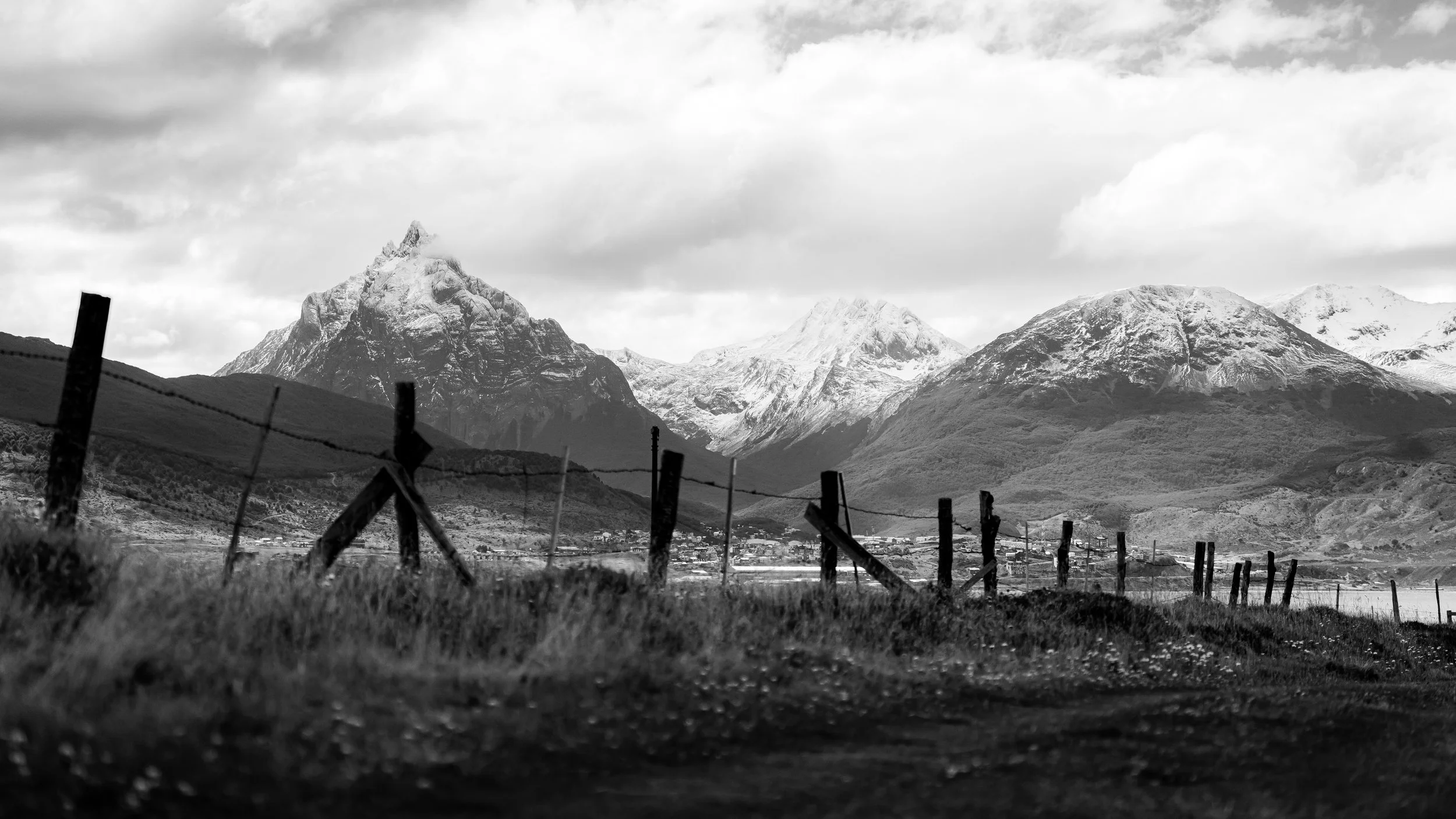Black and white photograph of rugged mountains with snow-capped peaks, a town at the base, and a barbed wire fence in the foreground.