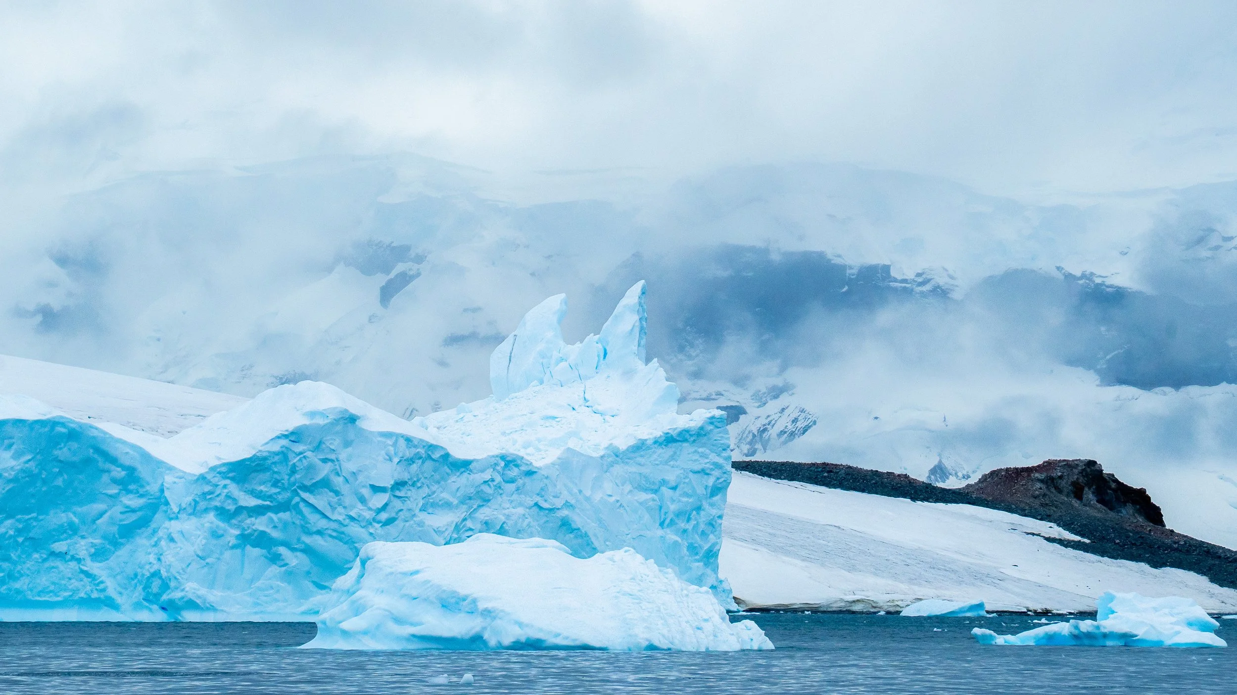 Iceberg floating in the ocean with a mountain and cloudy sky in the background.