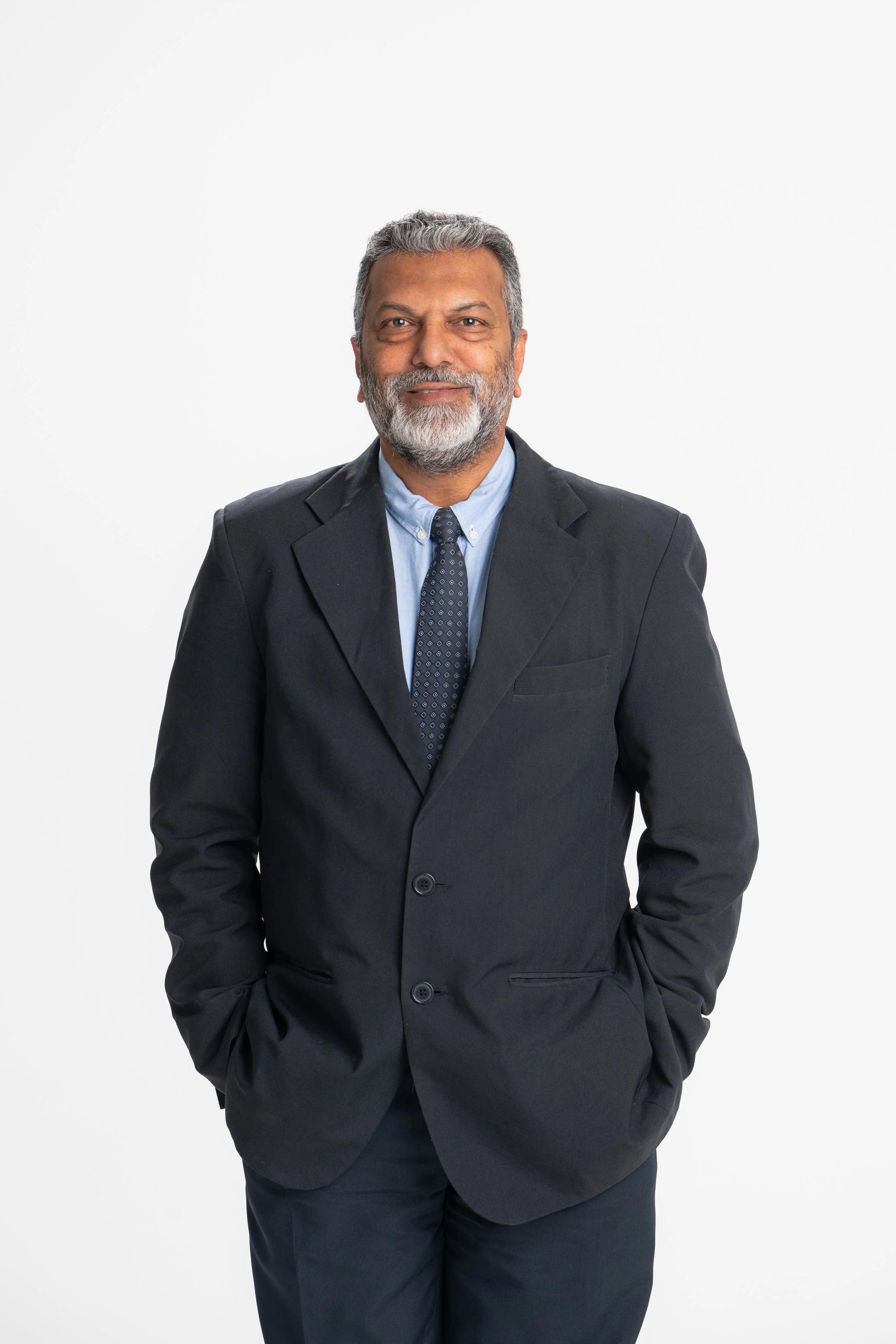 A middle-aged man with gray hair and a beard, wearing a dark suit, light blue dress shirt, and patterned tie, standing with hands in pockets against a plain white background.