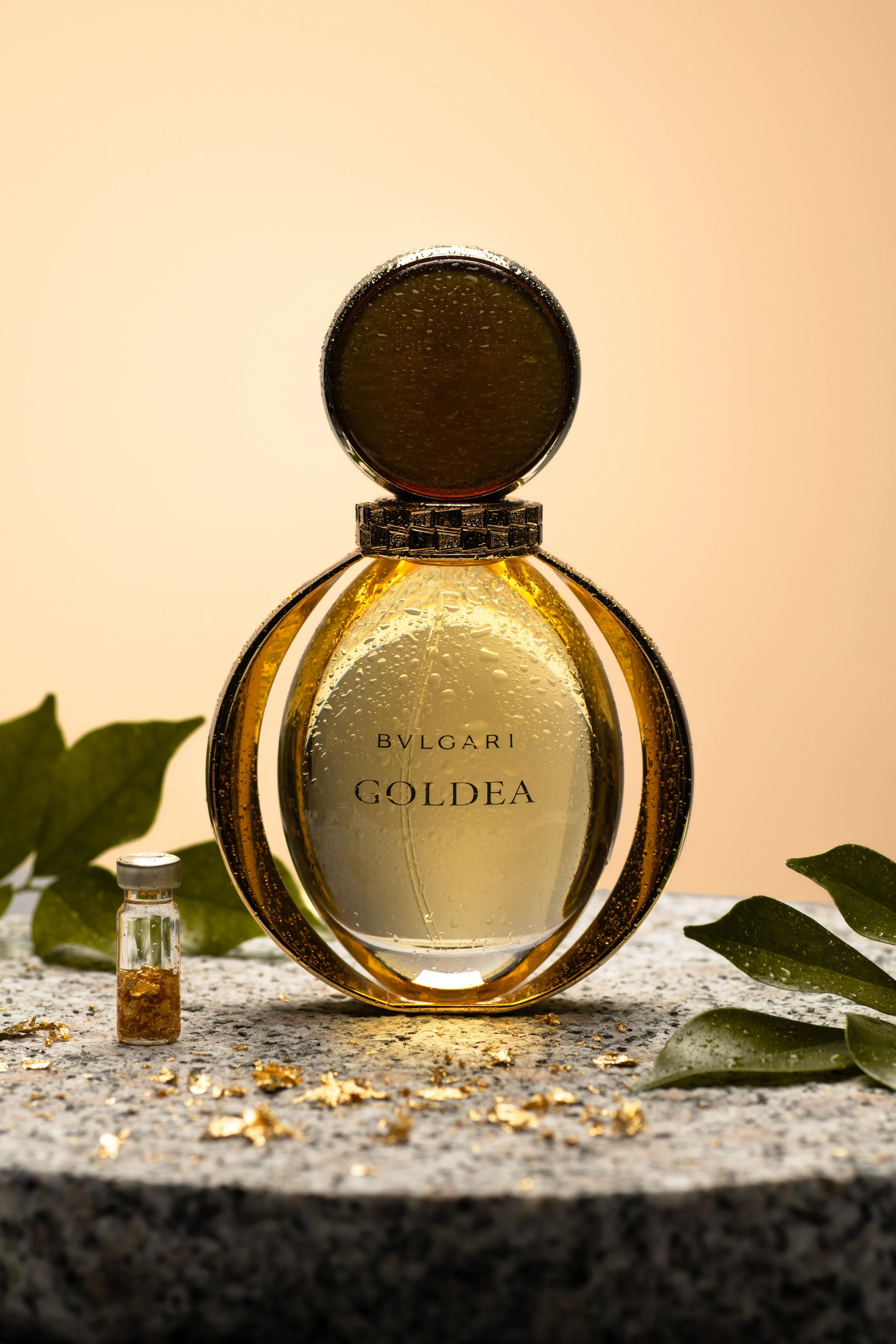 A triangular glass perfume bottle labeled Bvlgari Golden is surrounded by green leaves, a small vial, and scattered gold leaf flakes on a textured surface with a warm background.