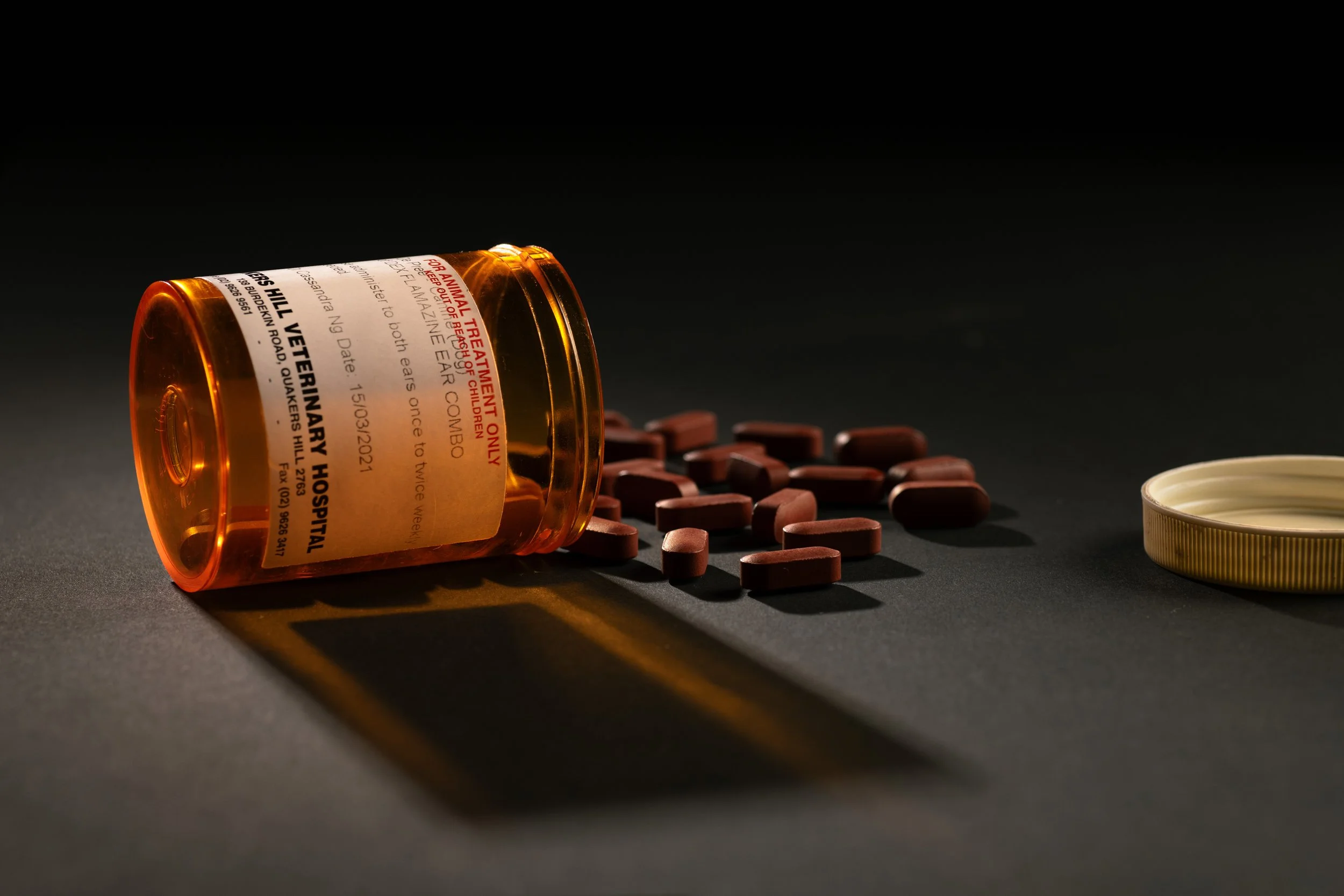 A spilled prescription pill bottle with red pills scattered around on a black surface, a white bottle cap is nearby.