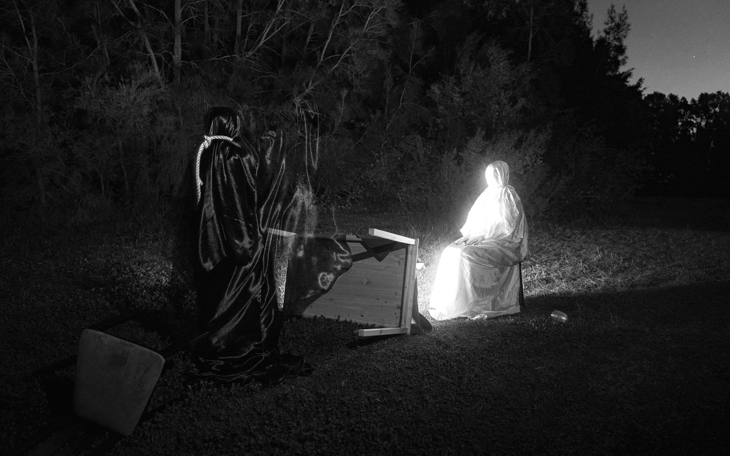 A nighttime outdoor scene with two people dressed in dark and white protective suits, possibly in a clandestine activity. One person is sitting on a chair near a bright light source, while the other stands nearby. There is a wooden box and some tools or equipment, with trees and night sky in the background.