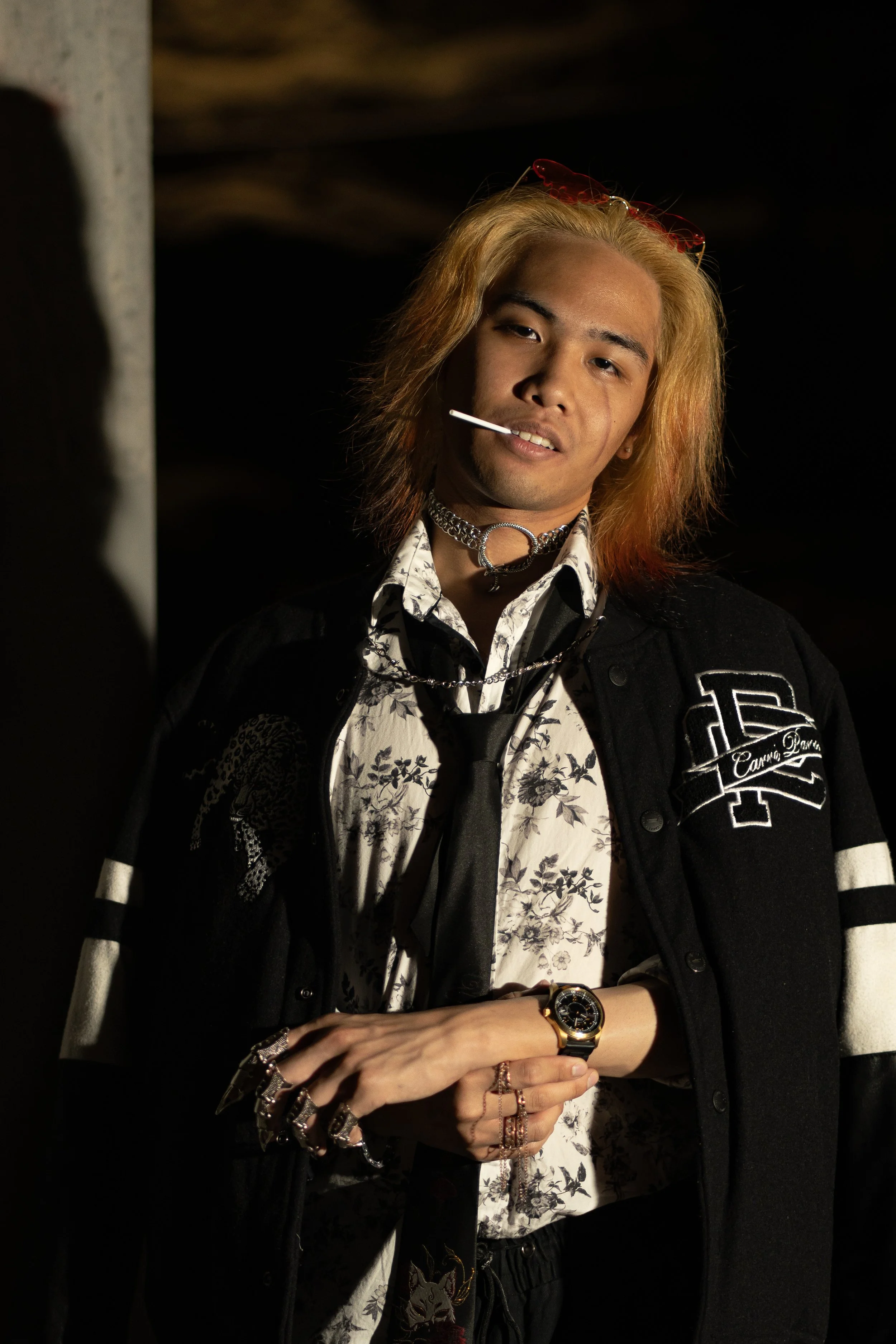 A young man with long, blonde hair wearing a varsity jacket, floral shirt, and accessories, standing against a black background.