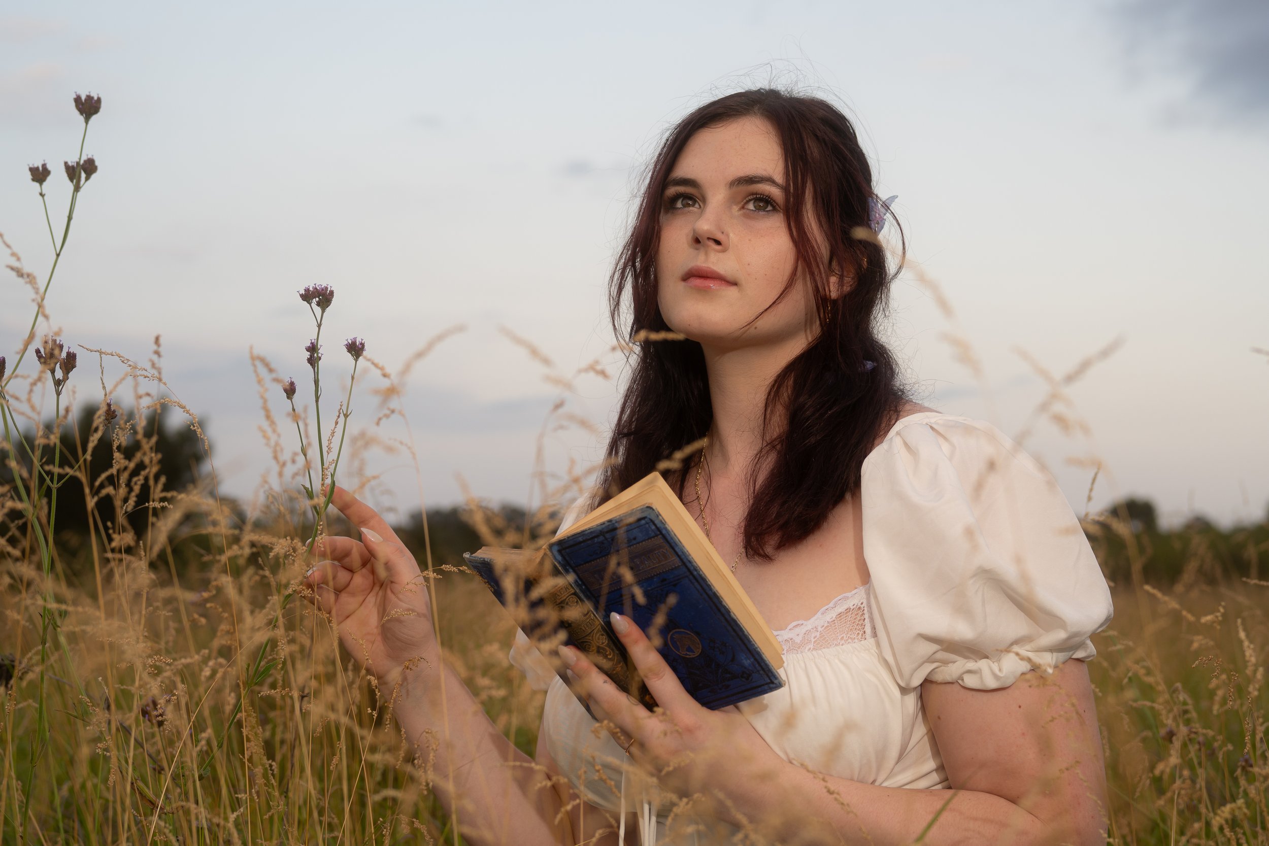 A woman with curly brown hair and a flower tucked behind her ear stands in a field of tall grasses and wildflowers, holding an open book and looking thoughtfully into the distance under a partly cloudy sky.