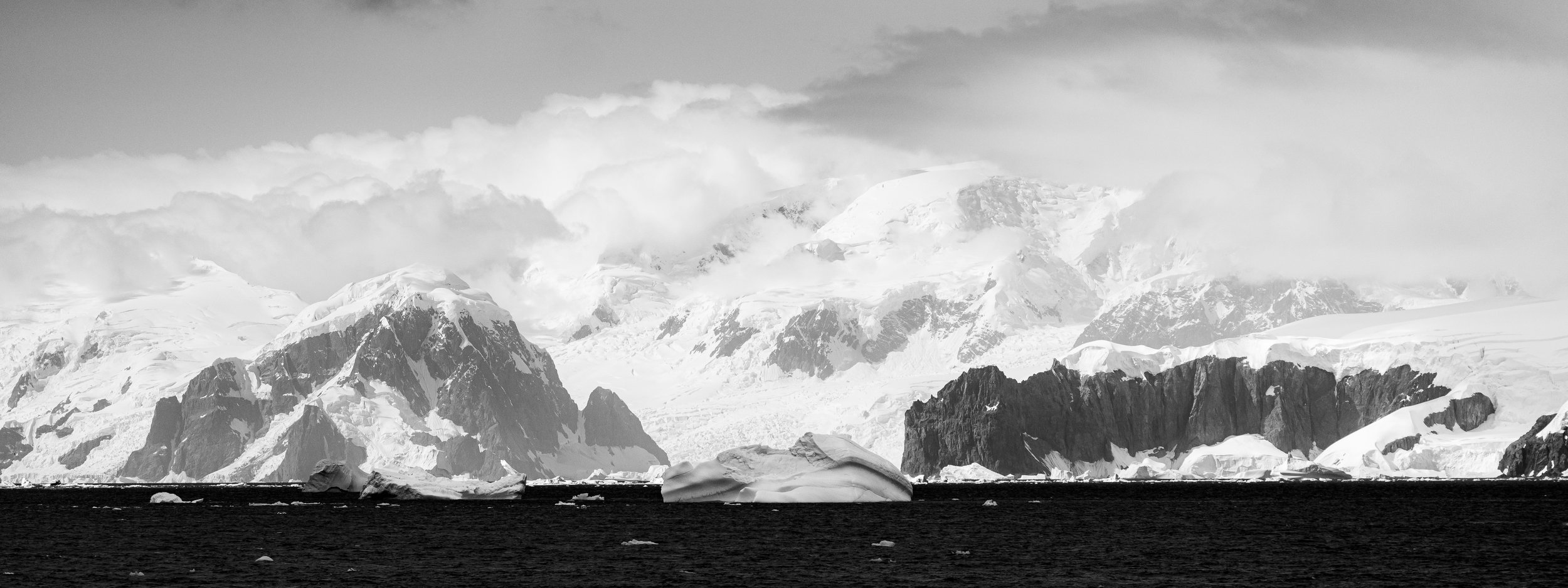 Black and white photograph of a large icy mountain landscape with snow-covered peaks in the background and an ocean with floating icebergs in the foreground.