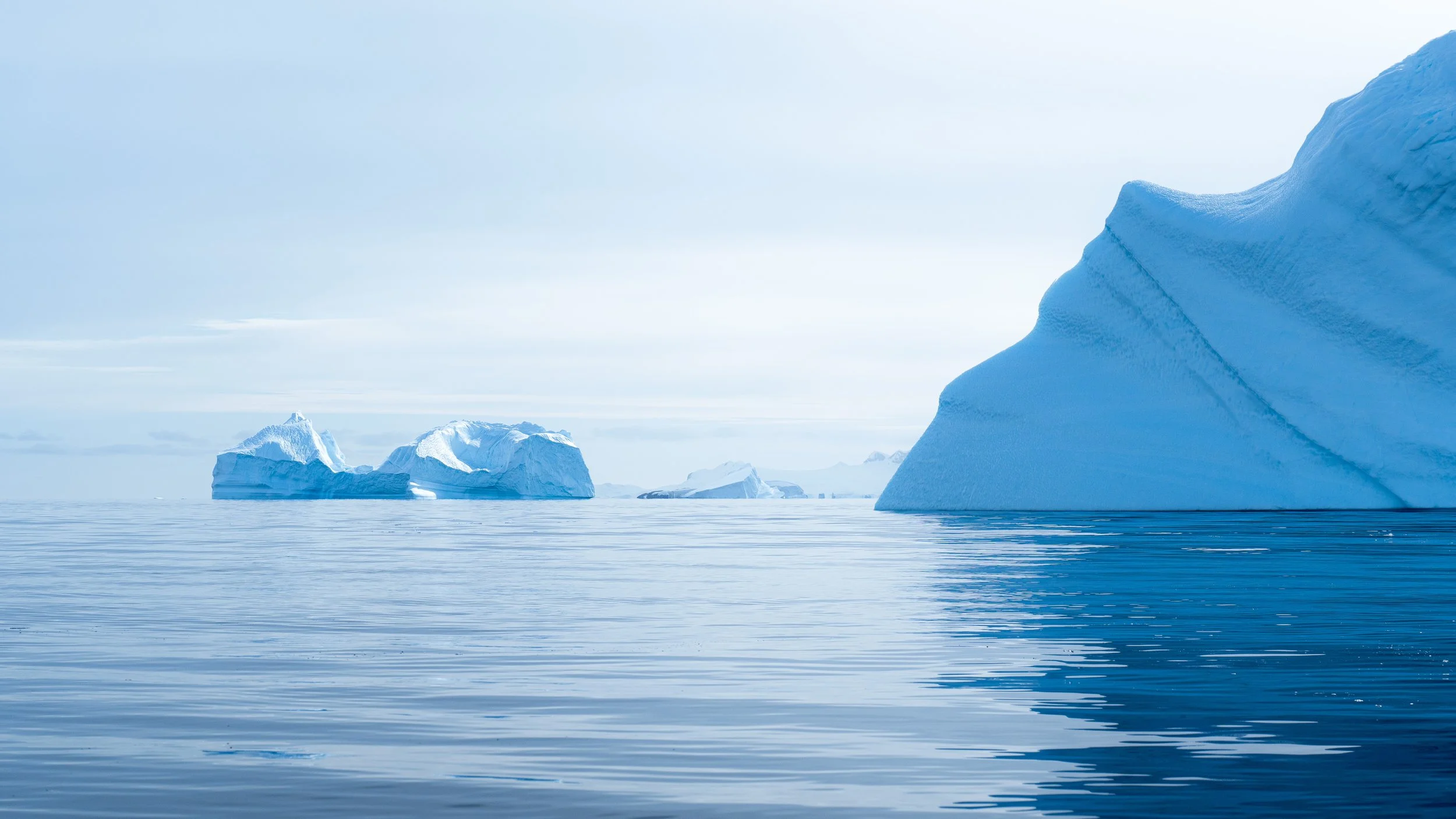 Calm ocean with large icebergs floating, including a prominent iceberg on the right side and several smaller ones in the background, under a cloudy sky.