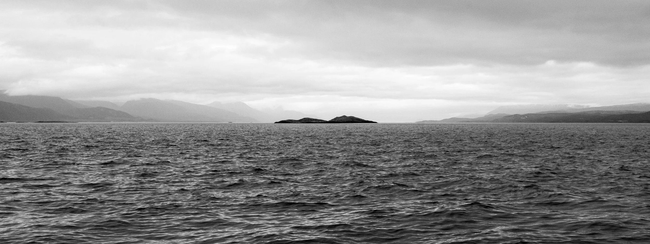 Black and white photo of a large body of water with small islands in the distance, mountains in the background, and a cloudy sky.