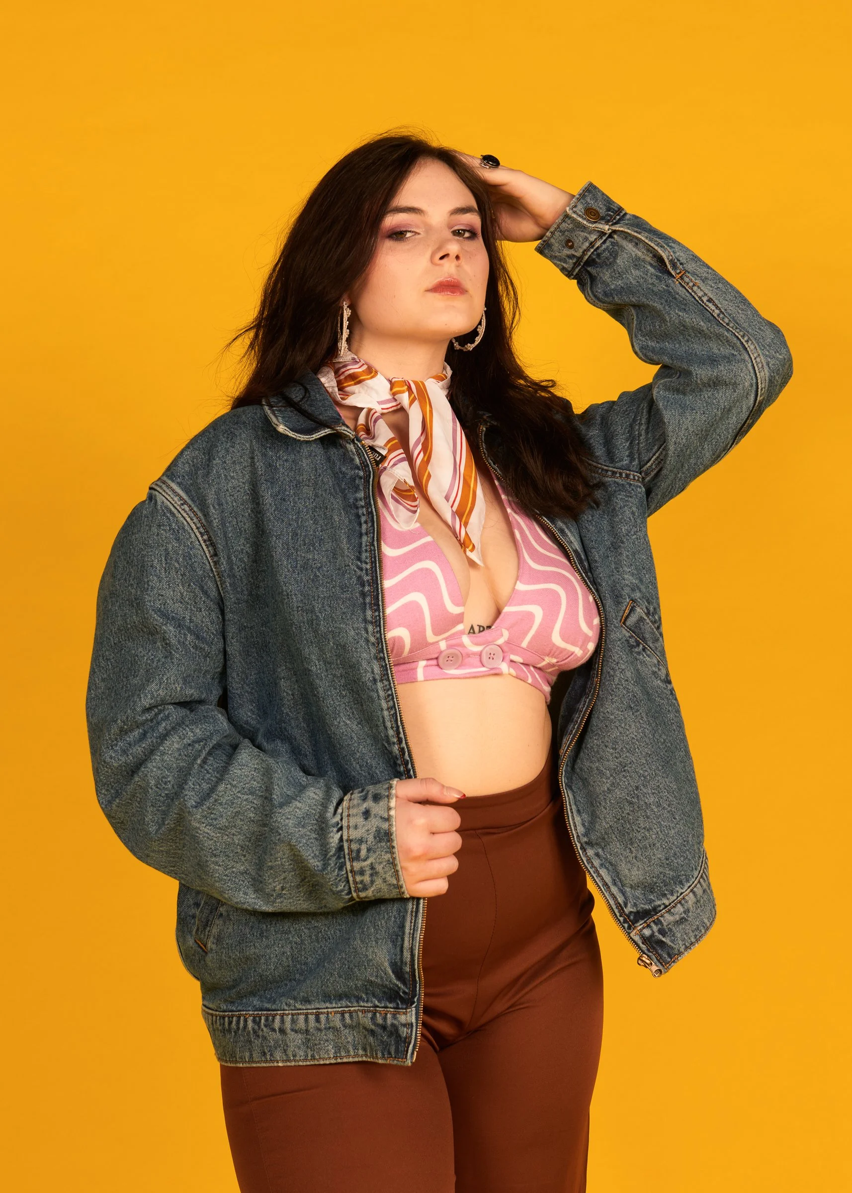 A young woman with long dark hair wears a denim jacket, pink patterned crop top, and brown high-waisted pants, posing against a yellow background.