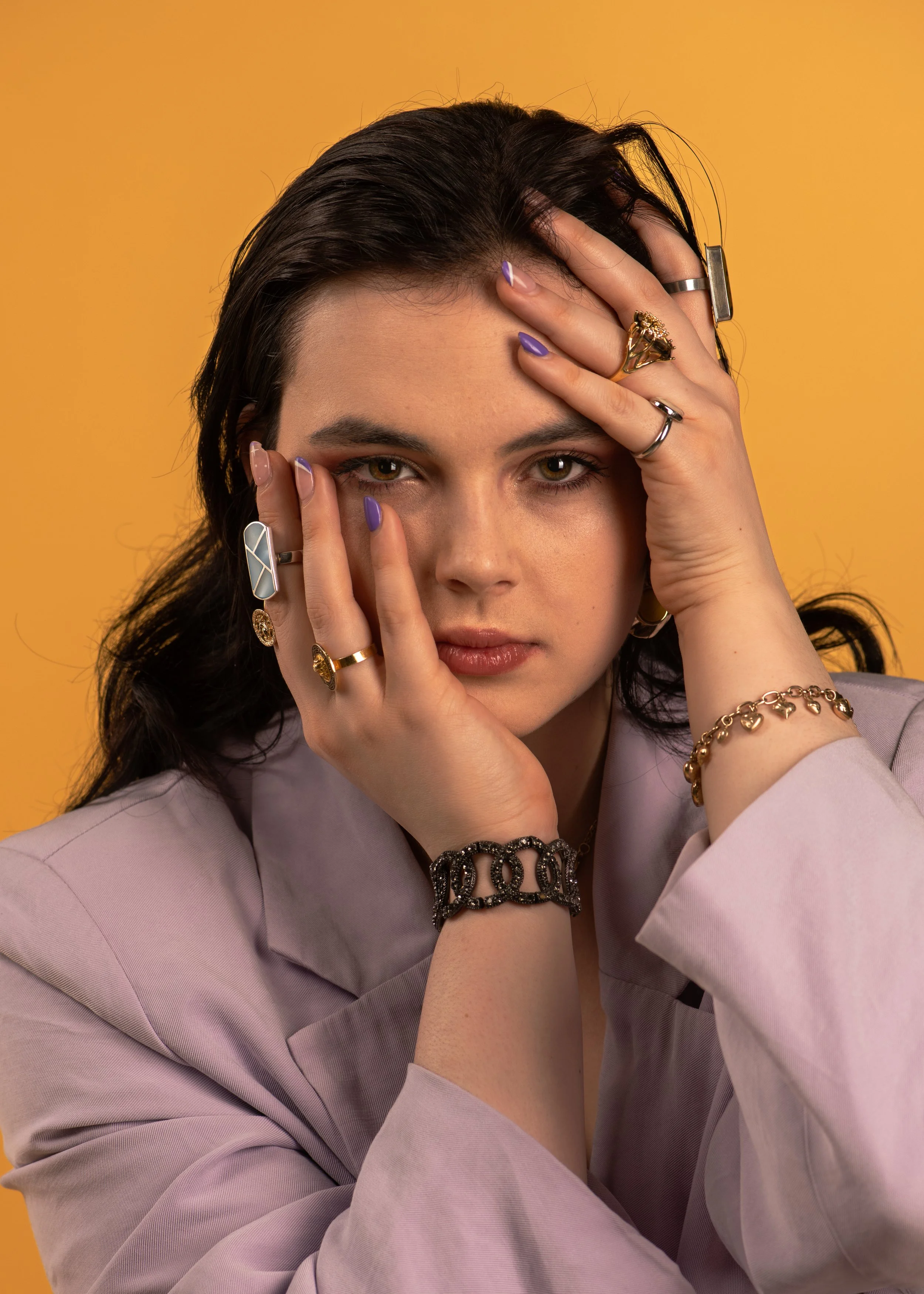 Portrait of a woman with dark, wet hair holding her face with both hands, wearing multiple rings and bracelets, against a yellow background.