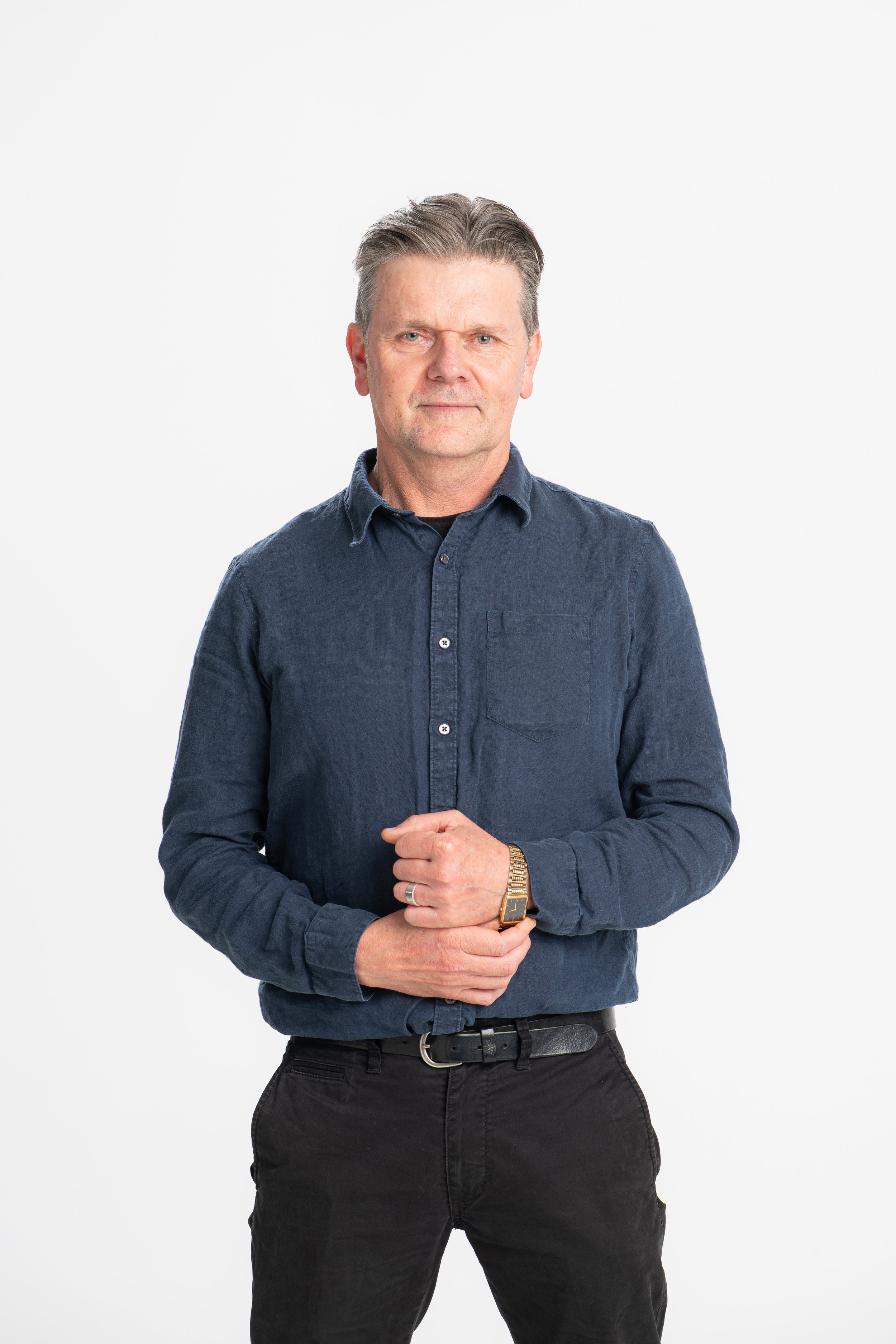 A middle-aged man with gray hair, wearing a dark blue denim shirt, standing against a plain white background. He has a serious expression, with hands clasped in front of him, and is wearing a watch on his left wrist.
