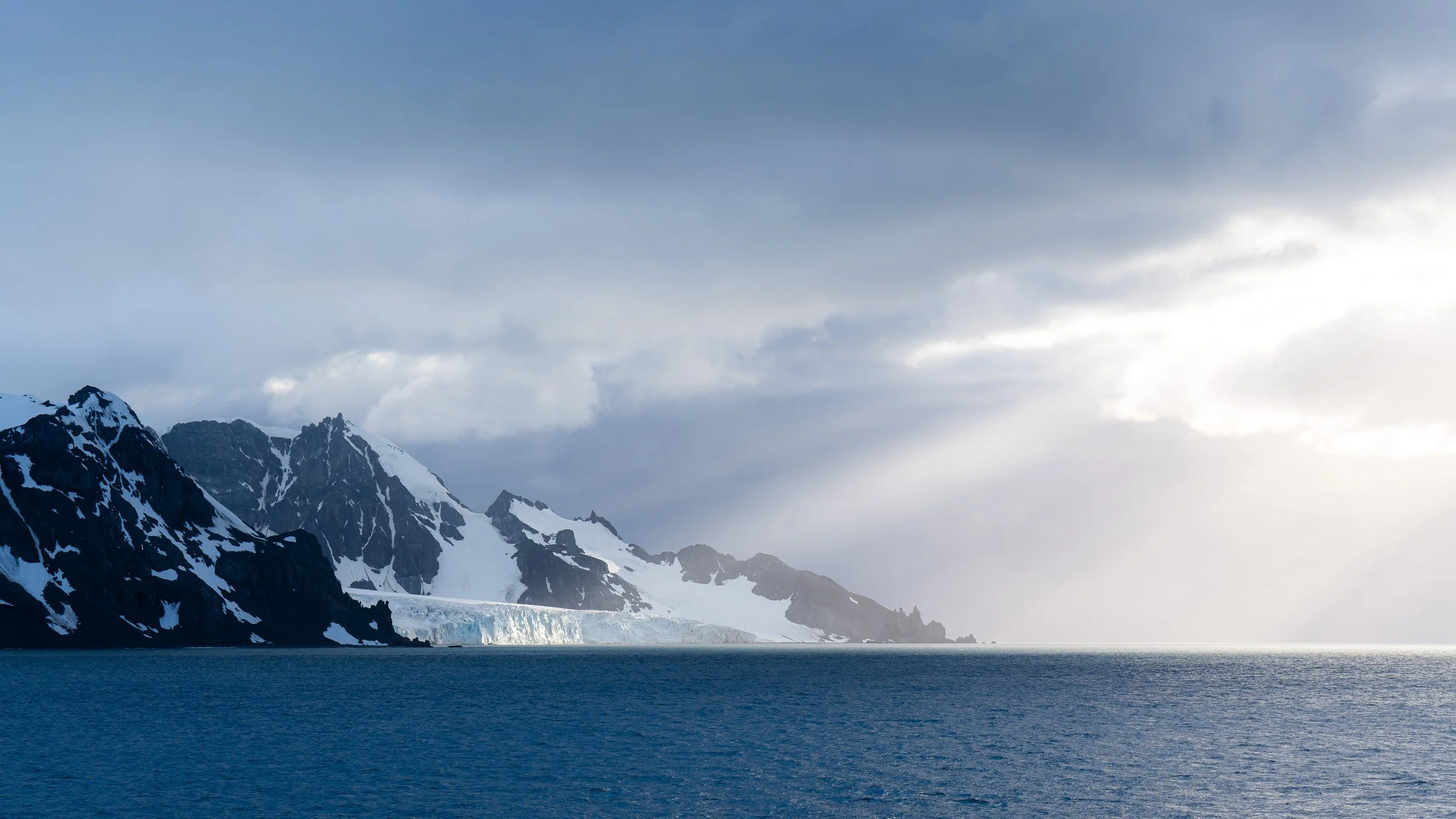 Snow-capped mountains with dark rocky peaks and icy glaciers, partially covered by clouds, over a body of water.
