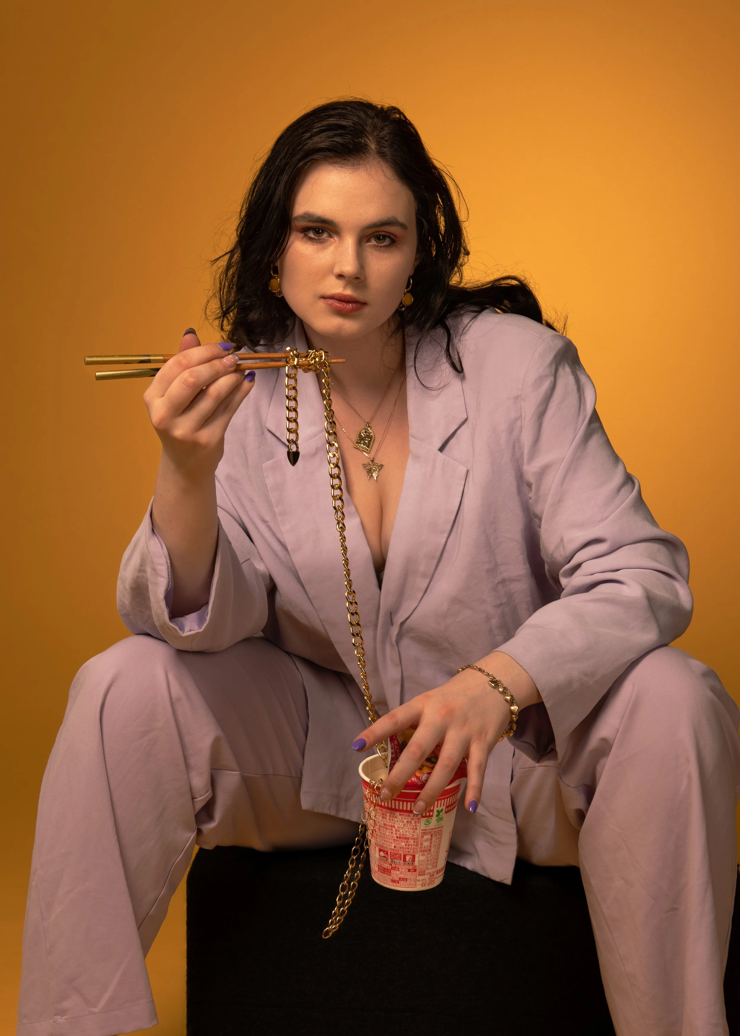 A woman with dark hair and light skin, wearing gold jewelry and a light purple blazer and pants, holds chopsticks near her face and sits against a yellow background. She is also holding a cup of instant noodles with a red and white label.