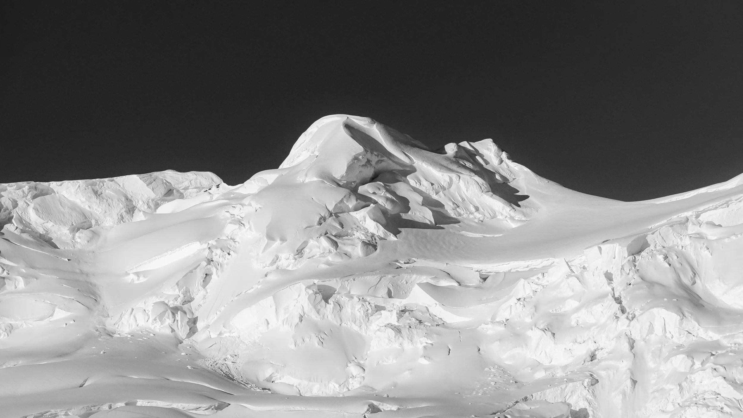 Black and white photo of snow-covered mountain with sharp peak against a dark sky.