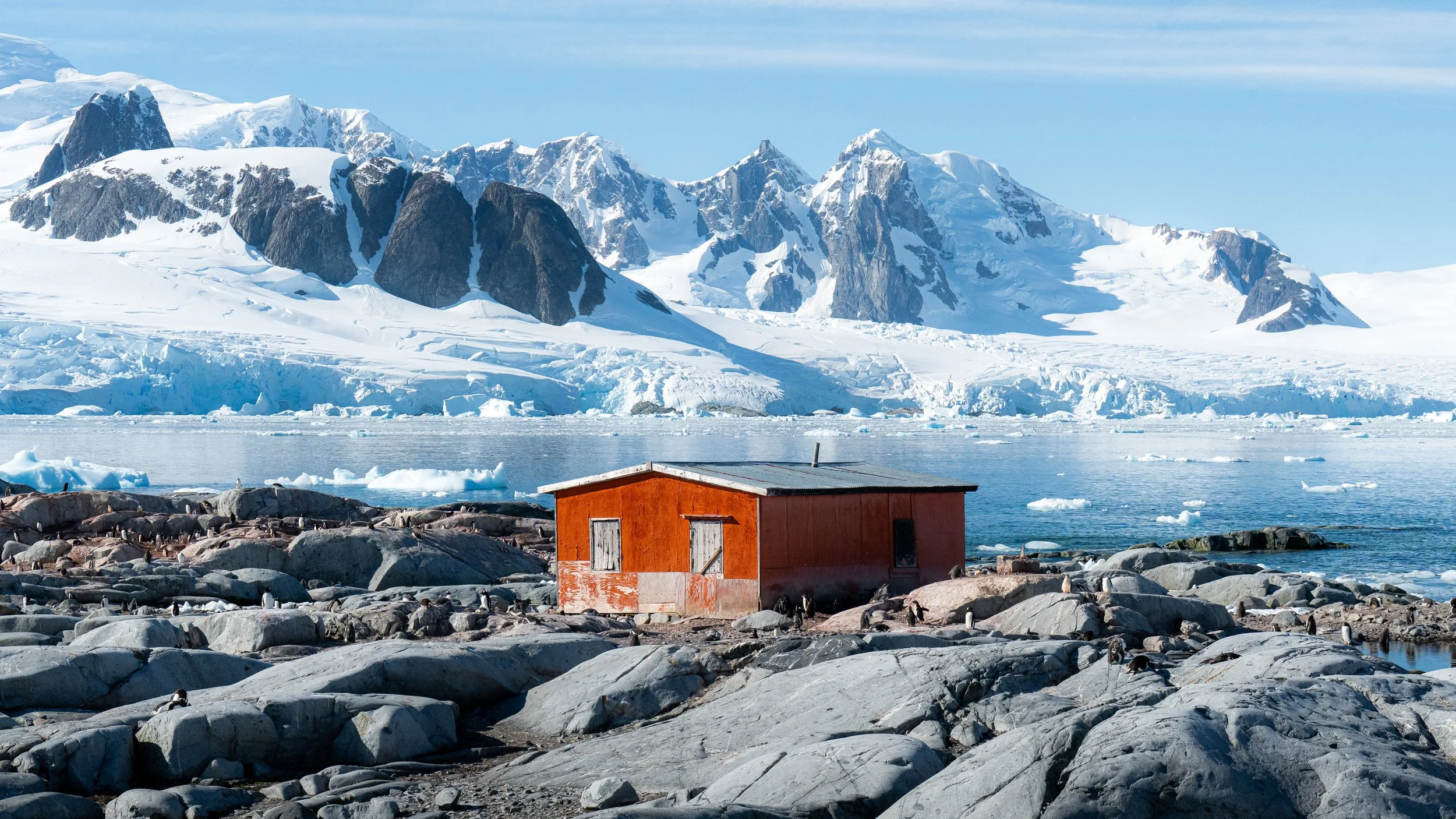 A small red house surrounded by rocky terrain and penguins on the shore of a frozen landscape with snow-covered mountains and glaciers in the background.