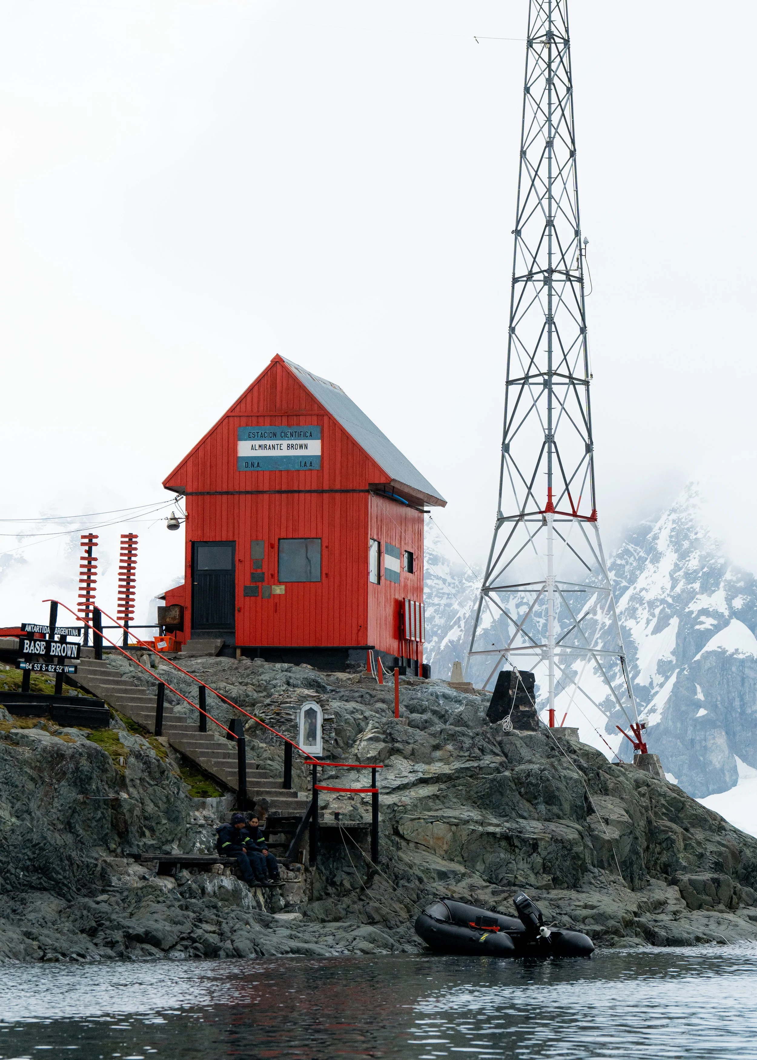 A red scientific station building with a sign reading "Estacion Cientifica Almirante Brown" located on a rocky shoreline in a cold, mountainous region. A large radio tower and some people sitting on rocks are visible near the water, with snow-covered