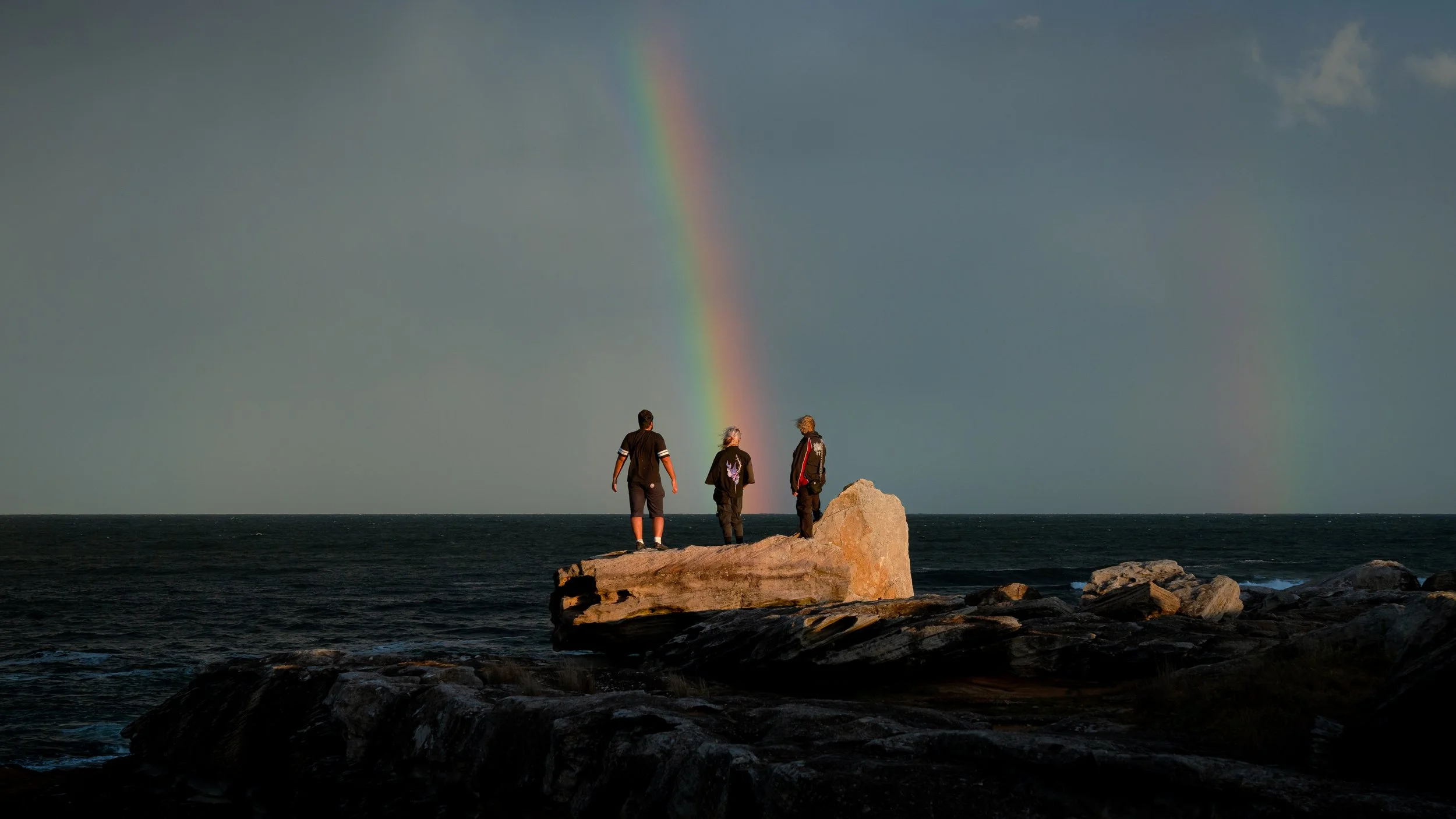 Three people standing on a rocky shoreline near the ocean, looking at a rainbow in the sky during a cloudy day.