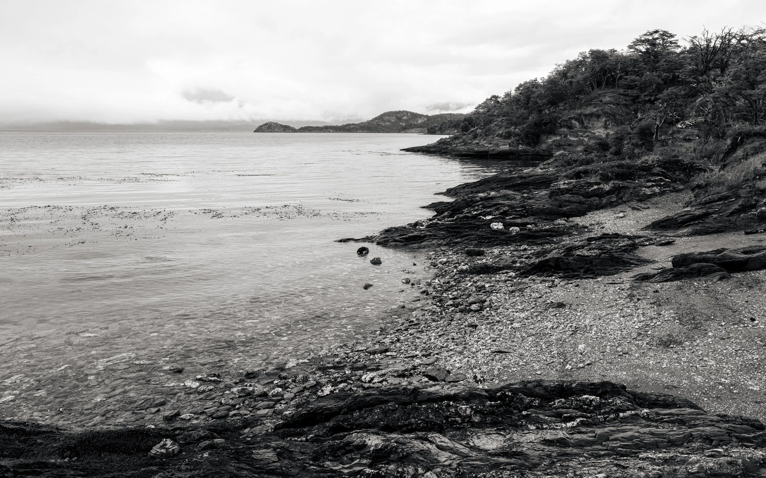 Black and white photo of a rocky shoreline with water and distant hills or islands under cloudy skies.