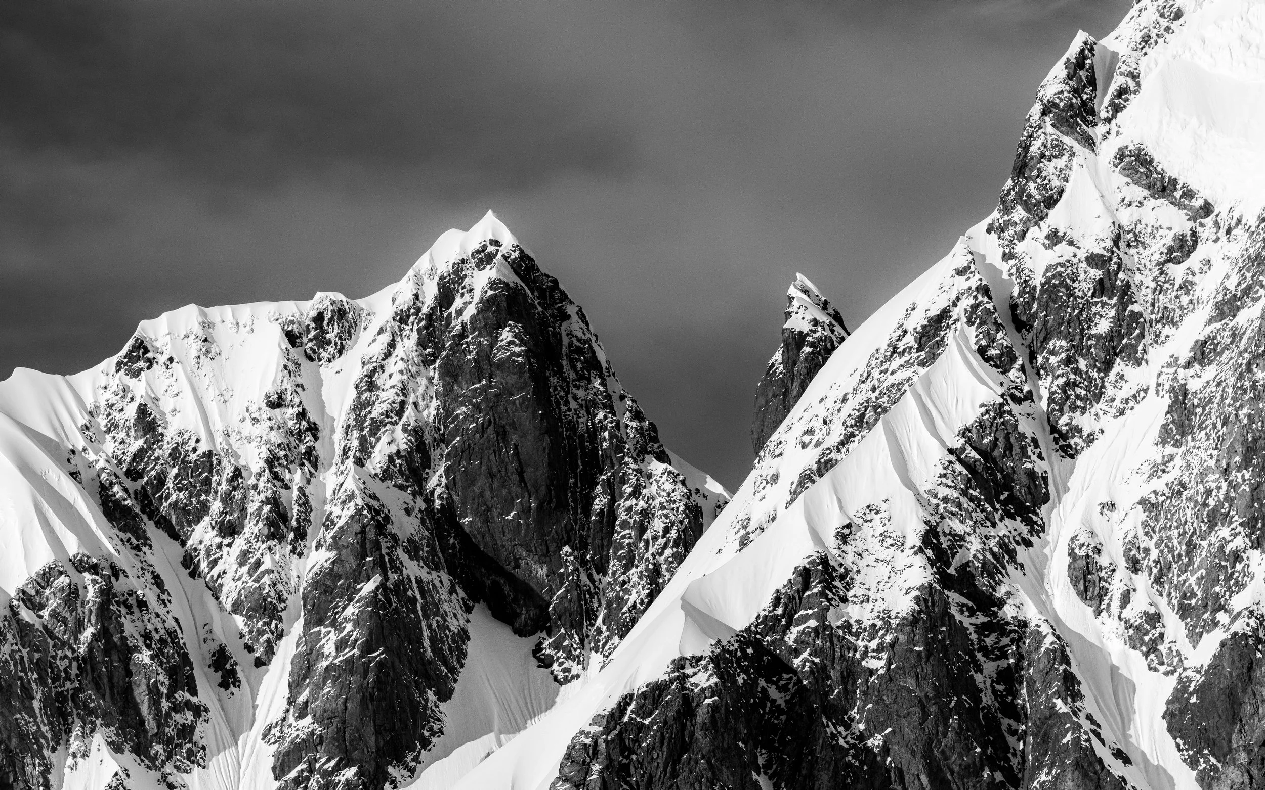 Black and white photograph of snow-covered mountain peaks with rugged, rocky faces and steep slopes under a cloudy sky.