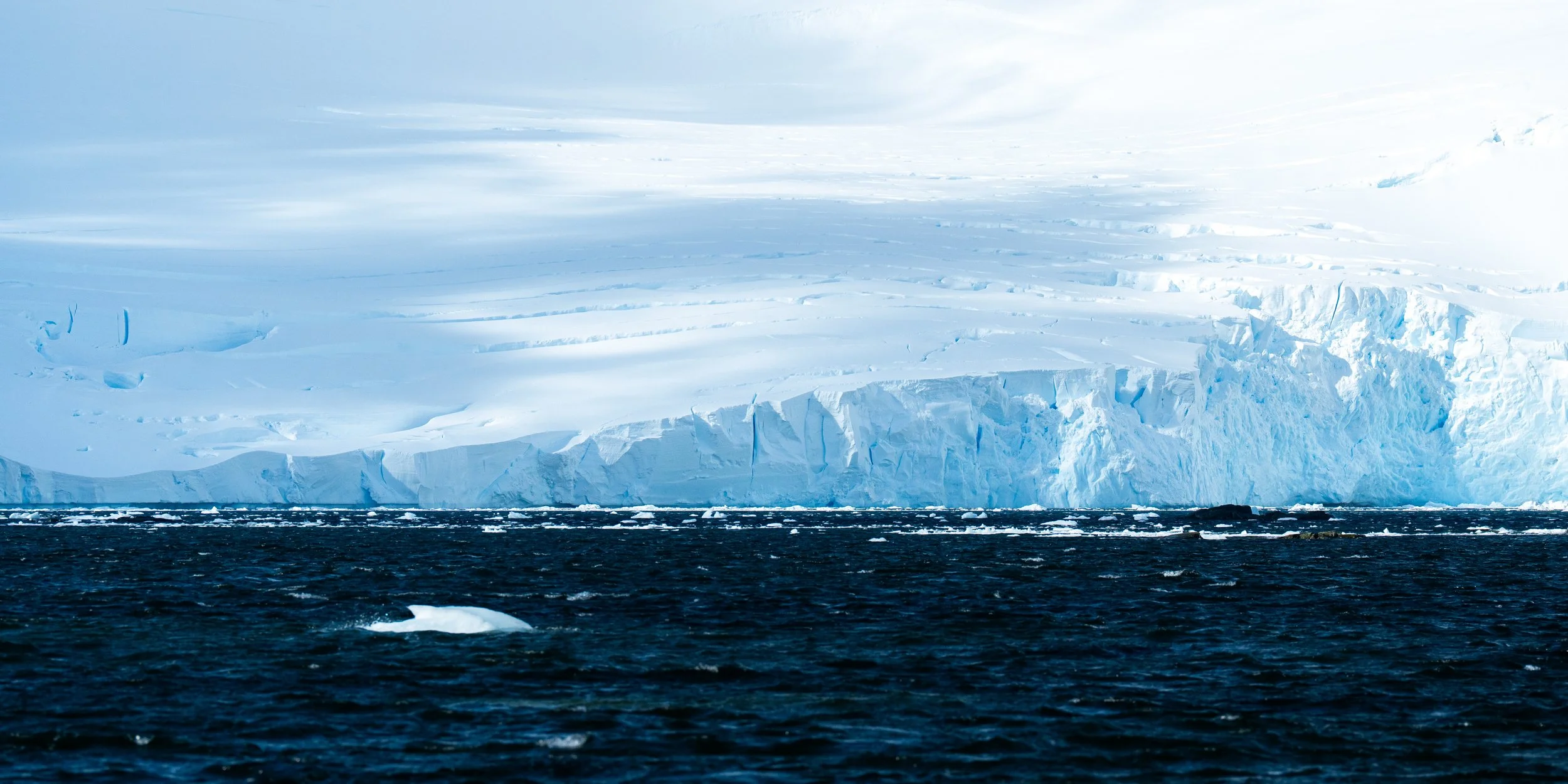 A large iceberg floating in the ocean with a clear sky and clouds above.