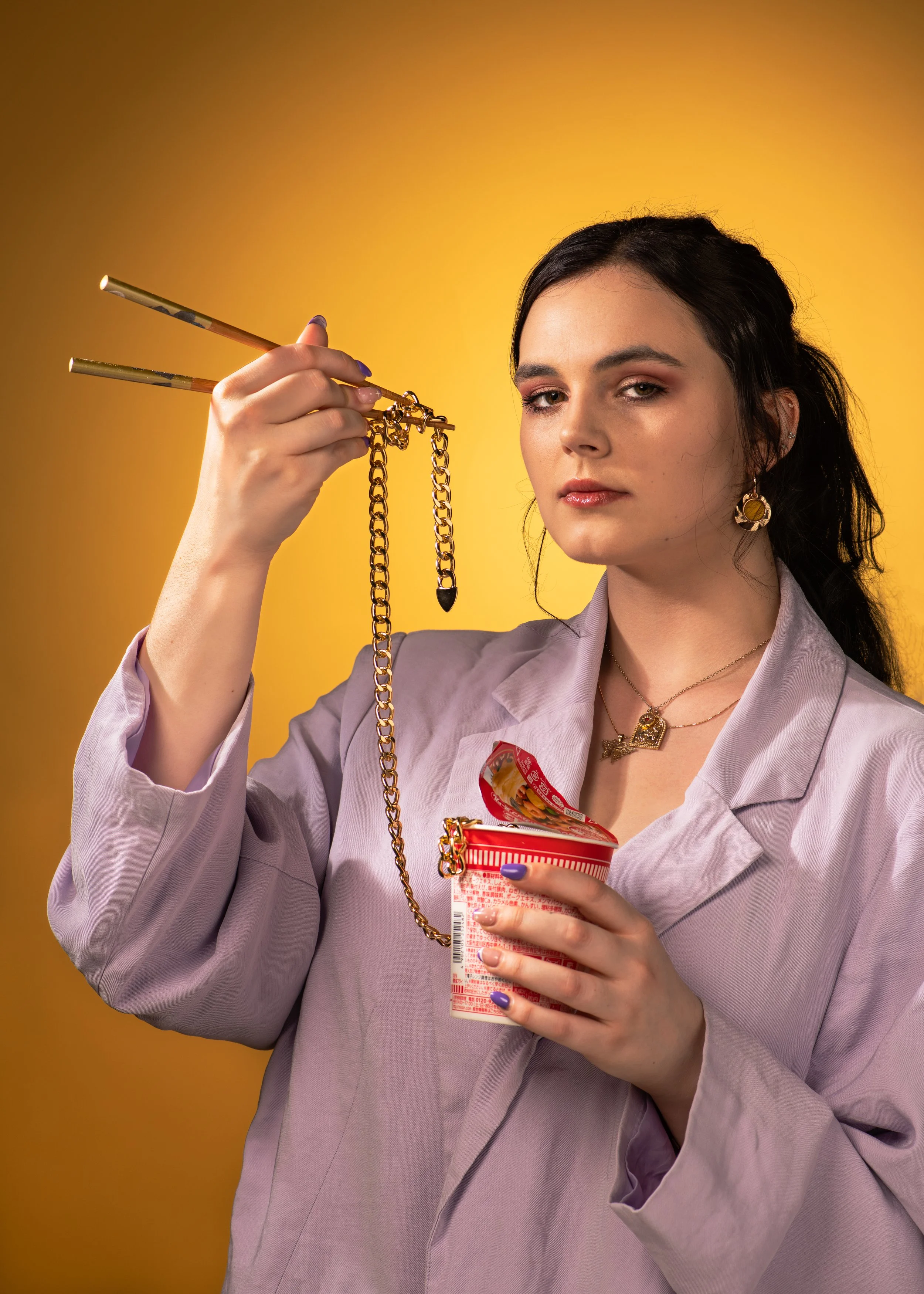 A young woman with dark hair and earrings, wearing a light purple blazer, holds a gold chain with charms in one hand and a cup of instant noodles in the other, against a yellow background.