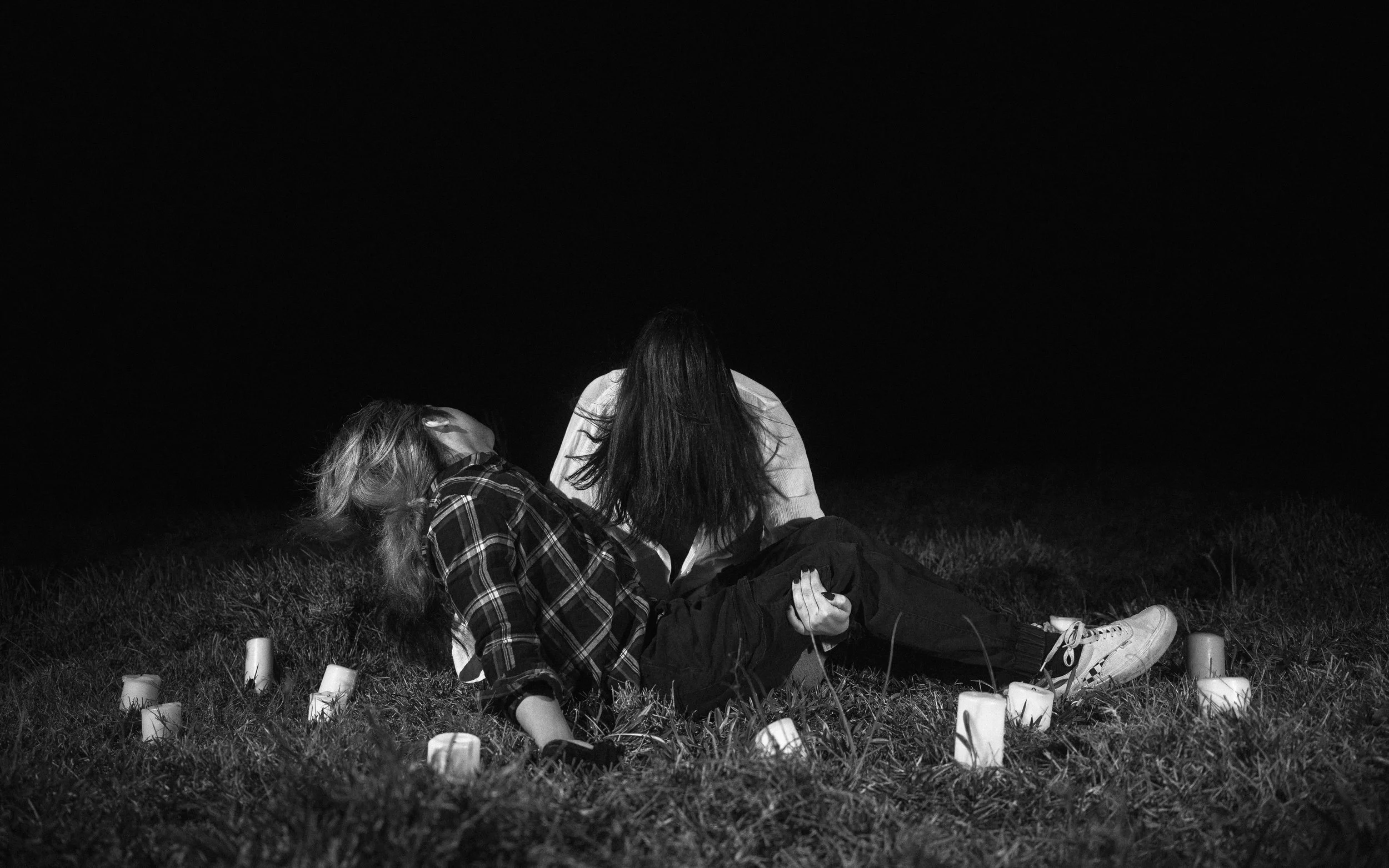 Two women sitting on the grass at night, surrounded by small candles, with a dark sky in the background.