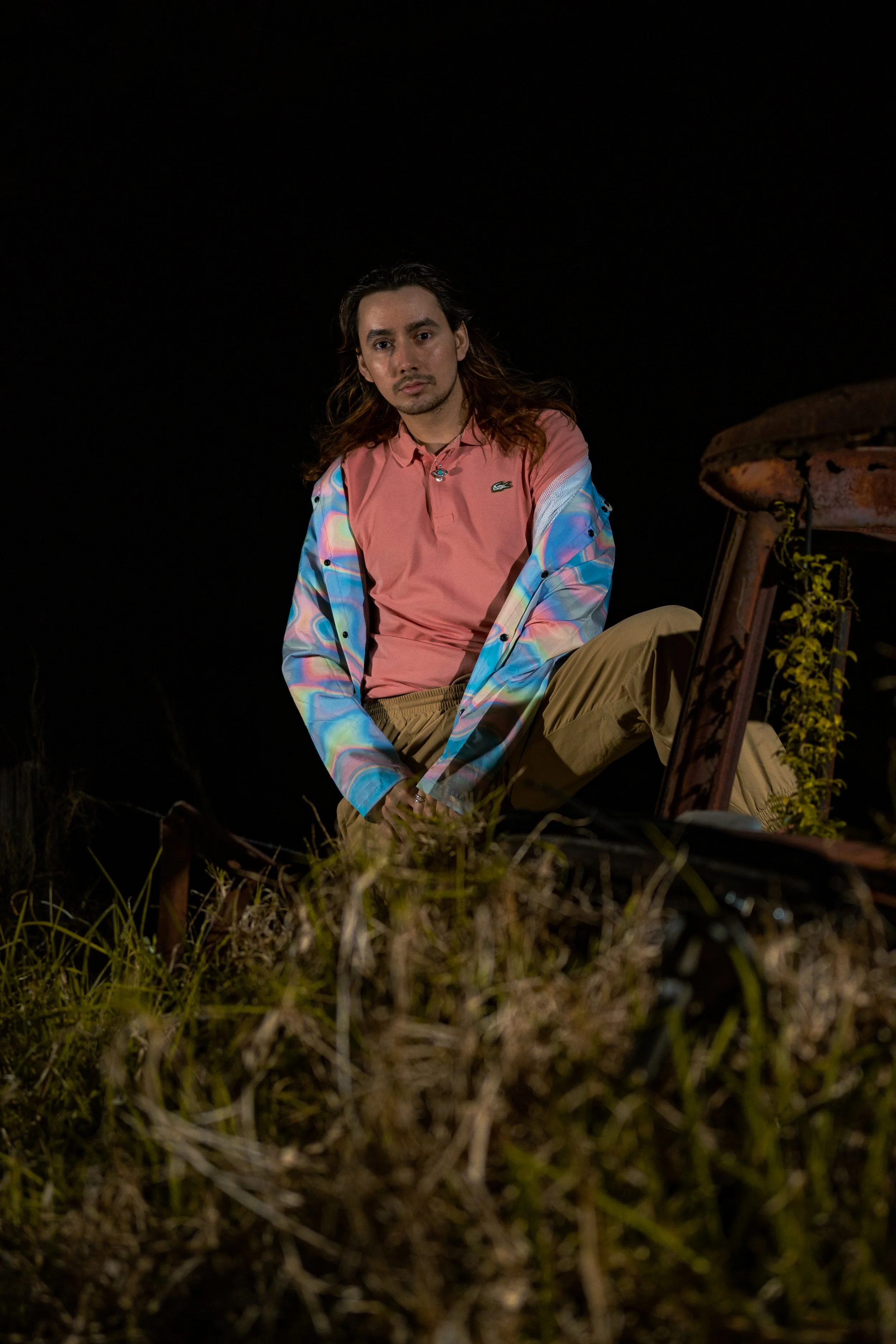 A man with long hair and a goatee, wearing a pink shirt, a colorful jacket, and beige pants, sitting next to an old rusted car at night with grass and plants in the foreground.