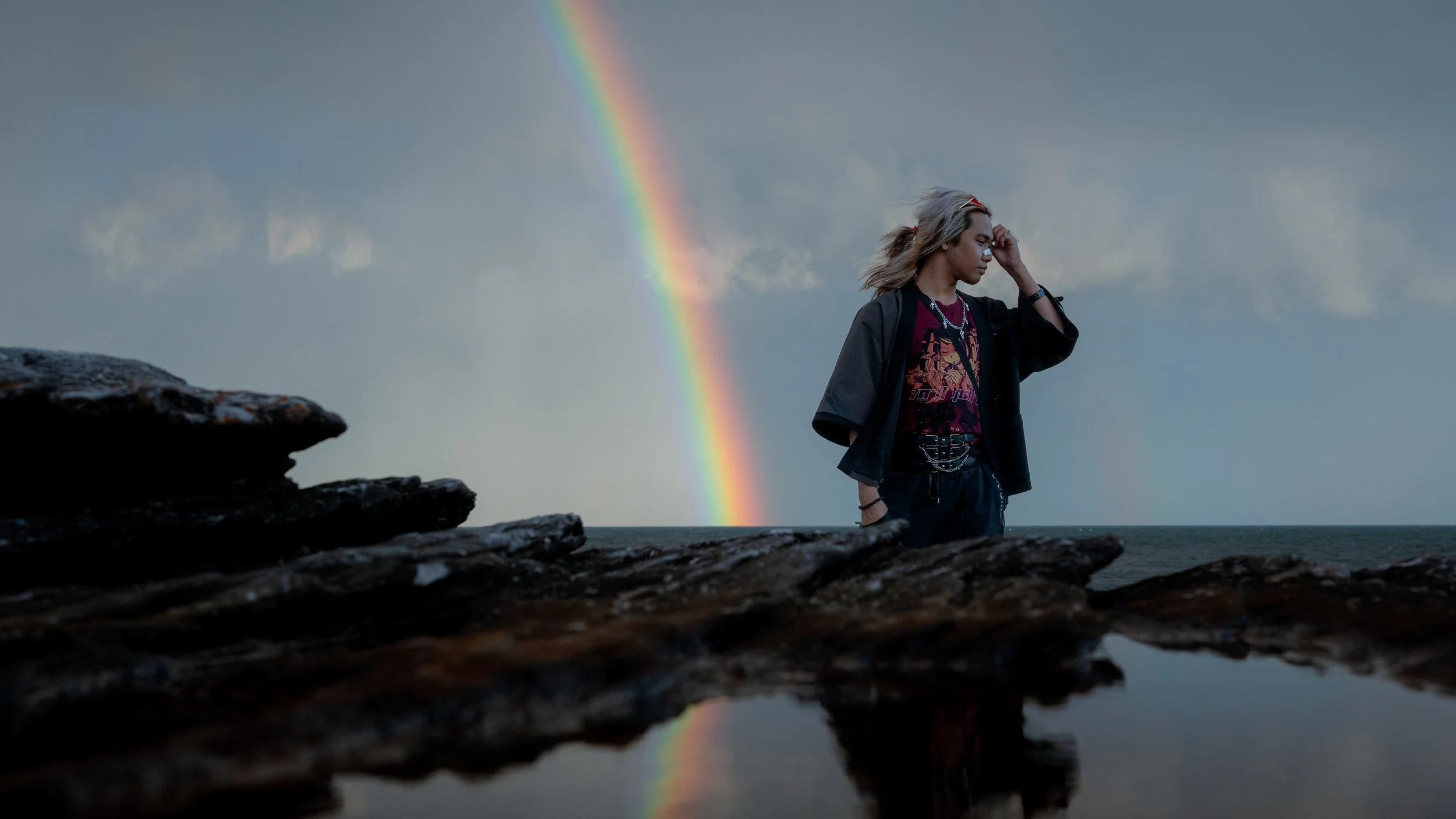 A young woman with blonde hair standing outdoors near a body of water during cloudy weather, with a rainbow visible in the sky behind her.