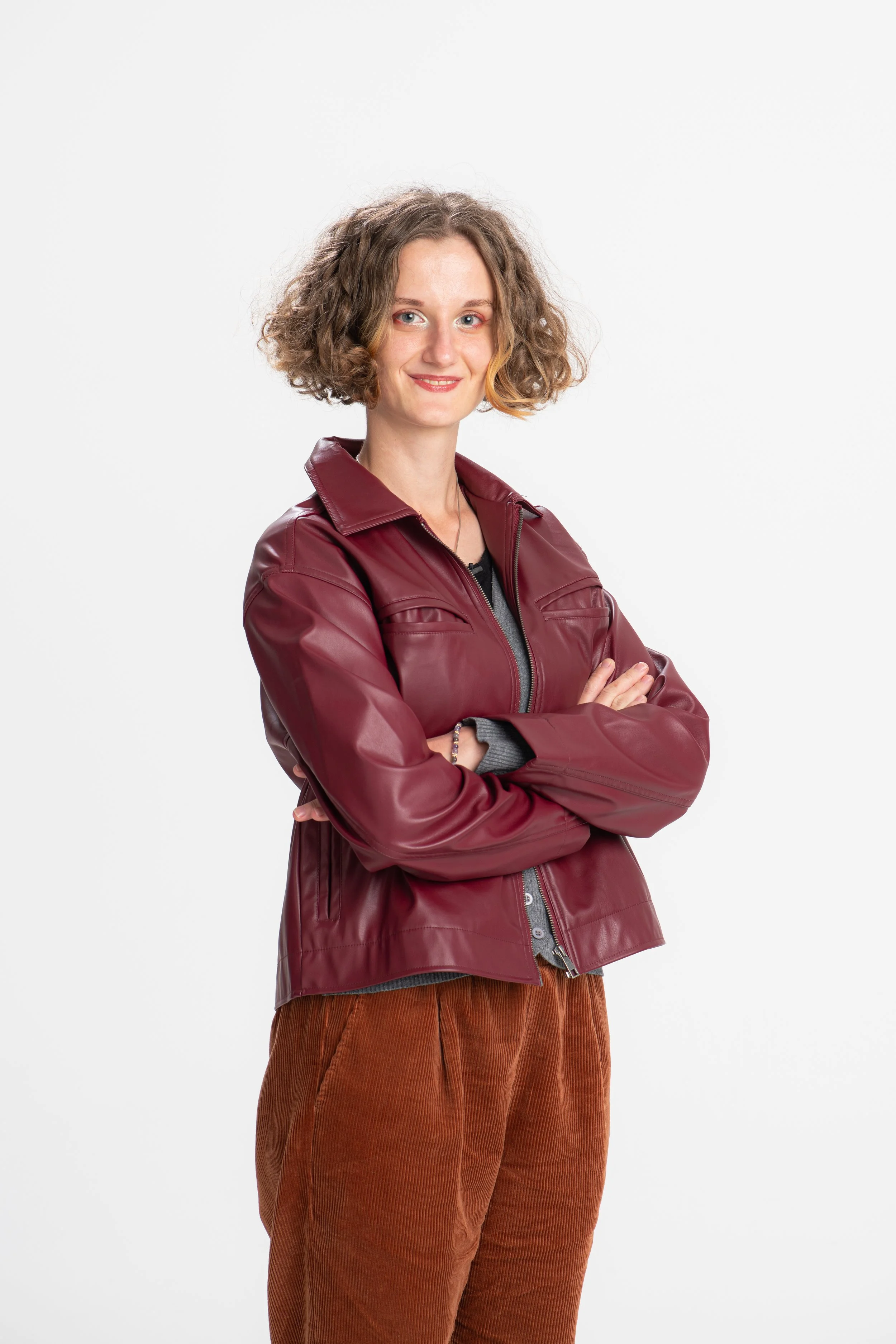 A woman with curly, shoulder-length hair wearing a burgundy leather jacket, a black top underneath, and brown corduroy pants standing against a white background.