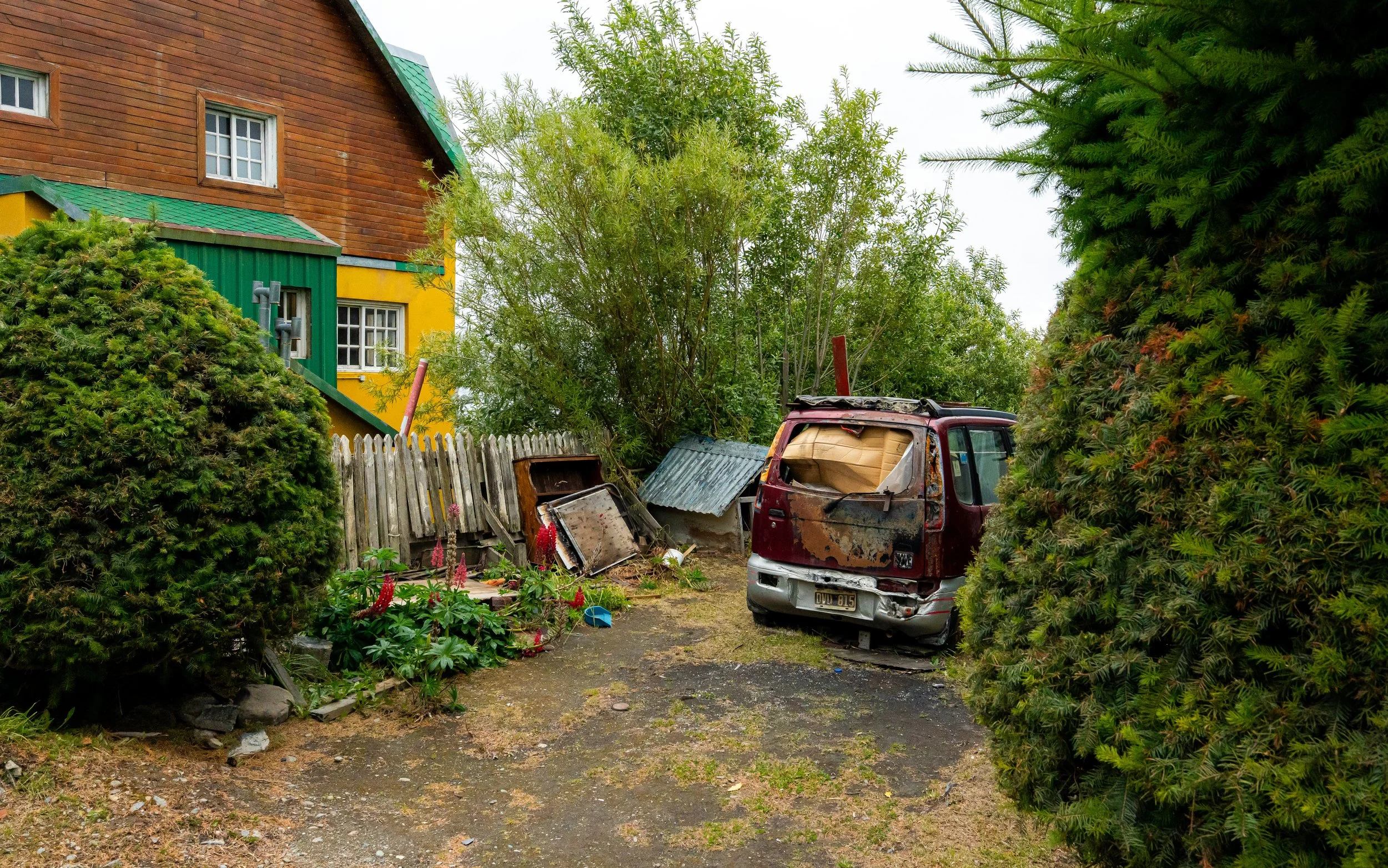A backyard scene with a wooden house painted yellow and green, surrounded by tall bushes and trees. An old, rusty, abandoned car is parked in the yard, and various outdoor items, including a wooden fence and garden plants, are visible.