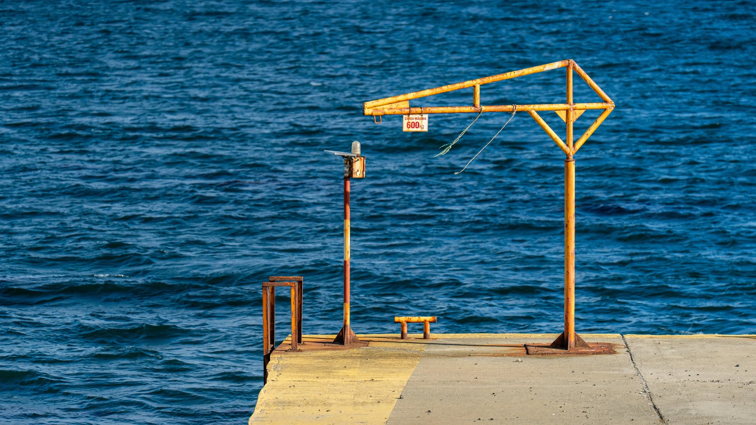 Rusty yellow metal structure on a concrete pier extending into the blue ocean water.