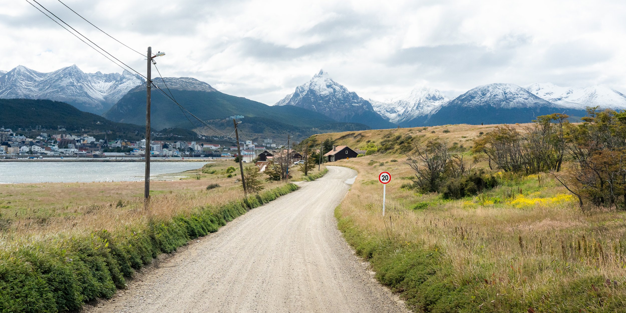 A dirt road winding through grassy fields with a 20 km/h speed limit sign, with snow-capped mountains in the background and a small town along the water under cloudy skies.