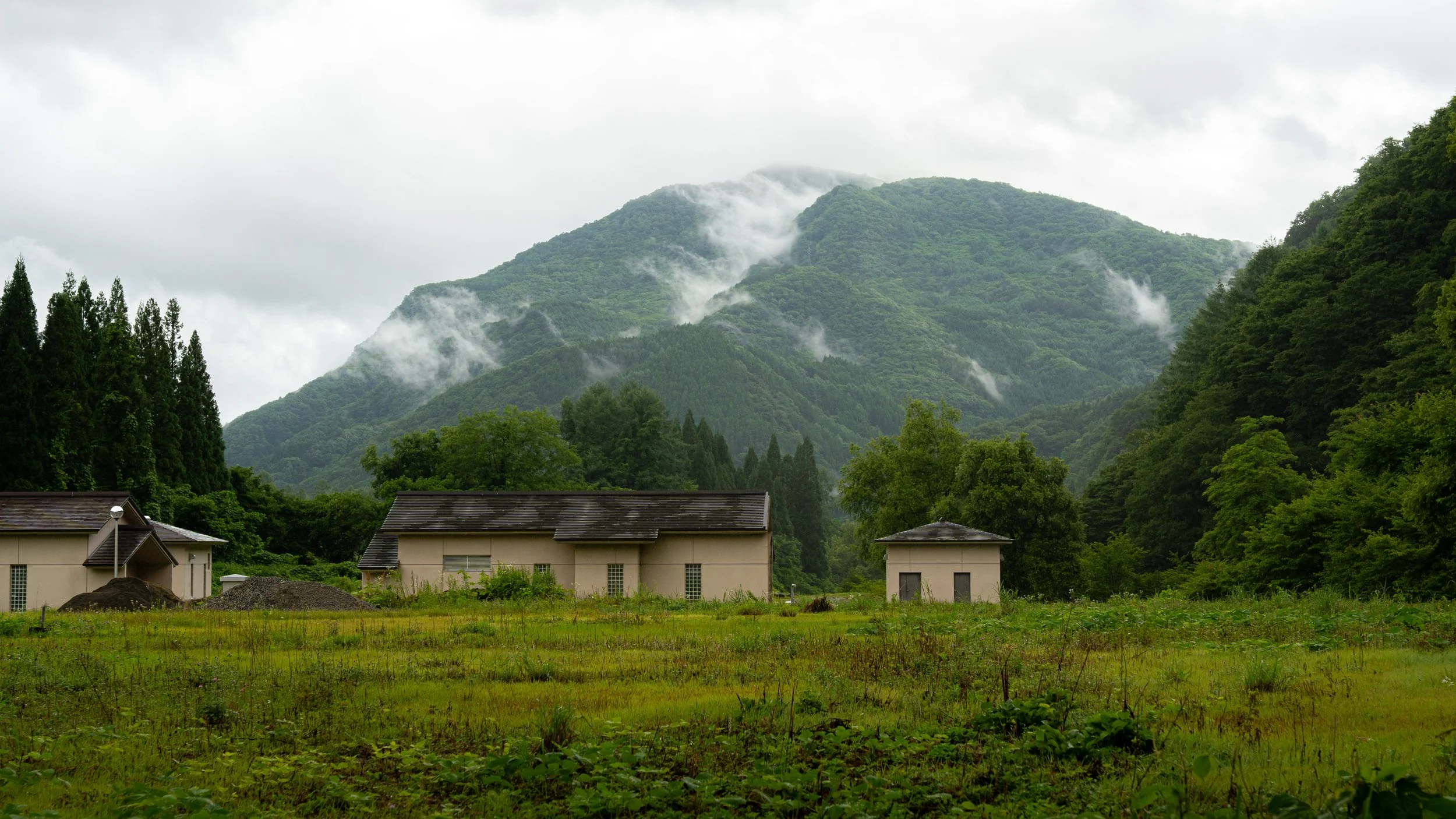 Green mountains with fog and clouds, some houses and a grassy field at the foreground.