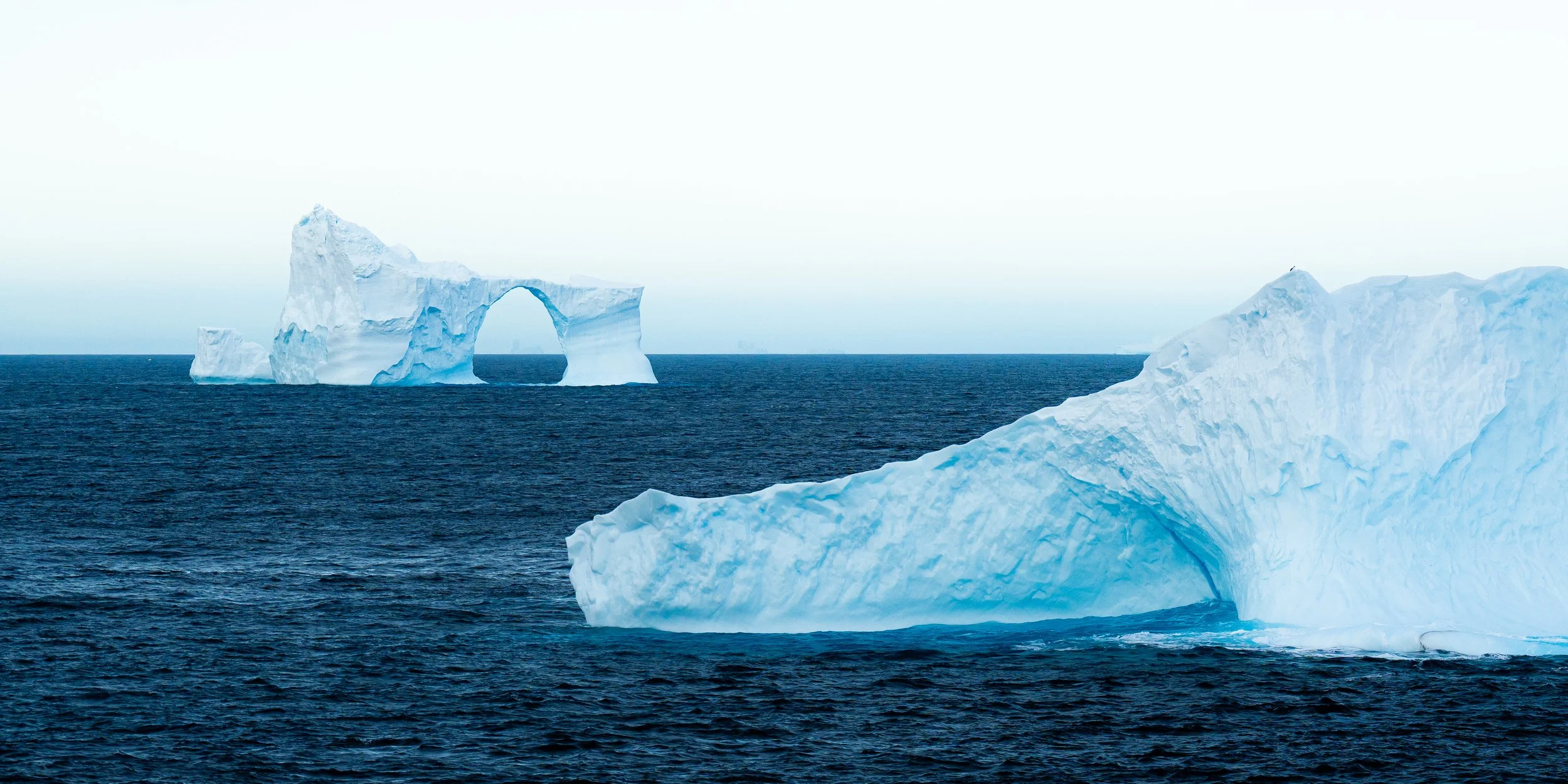 An ocean scene with large floating icebergs, including one with a natural arch, under a cloudy sky.