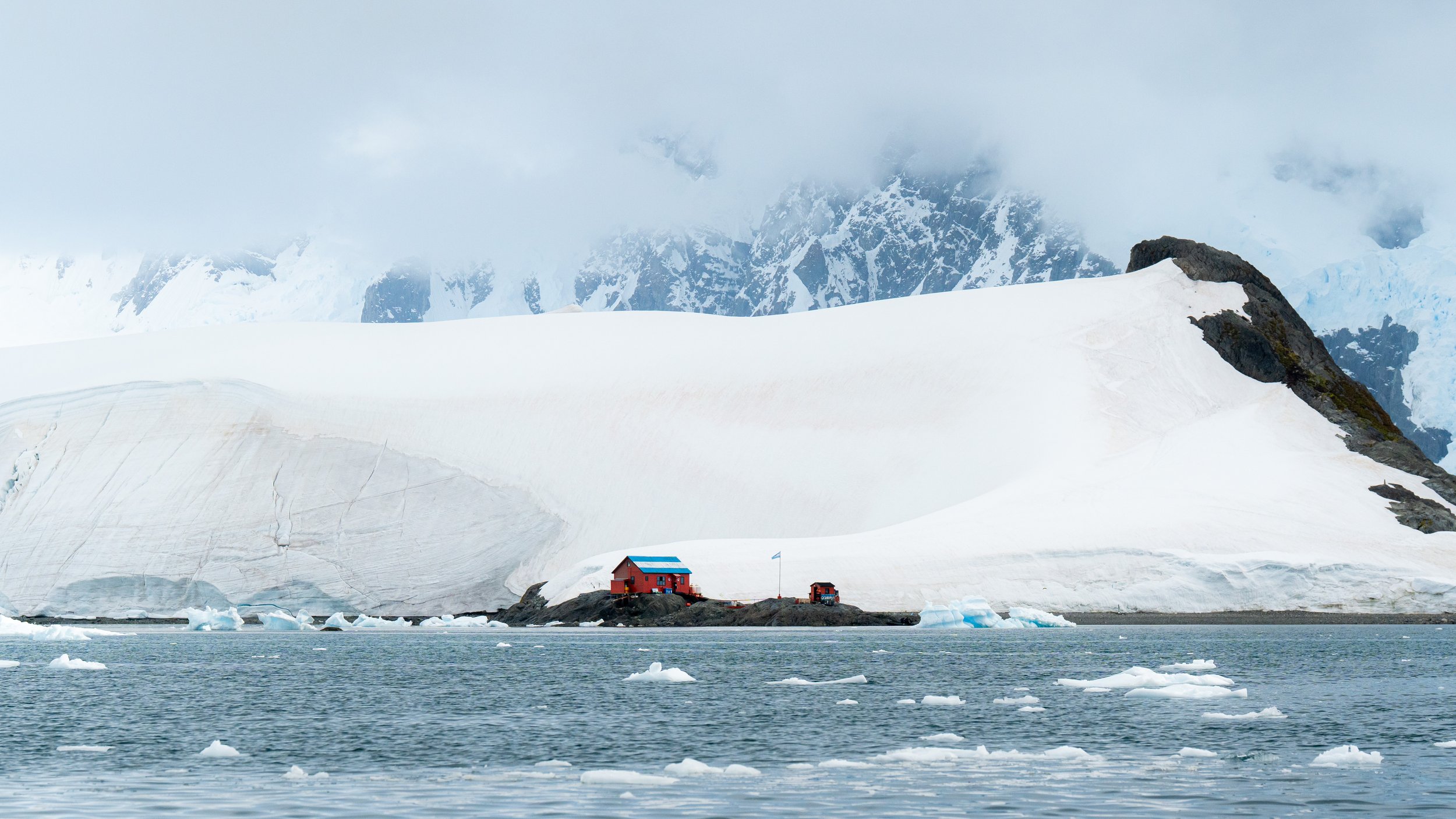 A red building located on a small rocky island in icy water, with large snow-covered mountains and glaciers in the background, partly shrouded in fog.