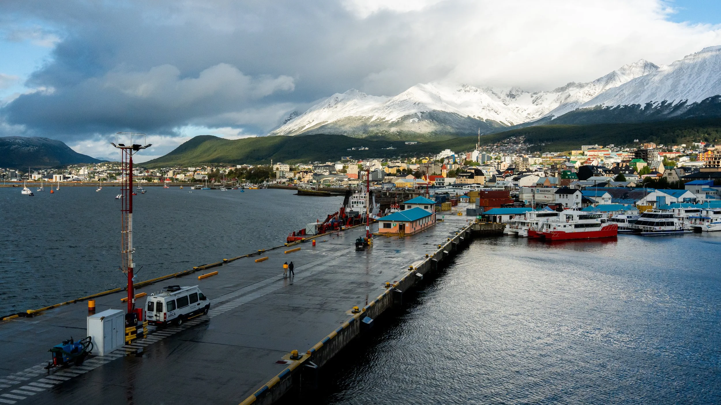 City port with boats docked, mountainous landscape in the background, cloudy sky, and a dock area with vehicles and equipment.