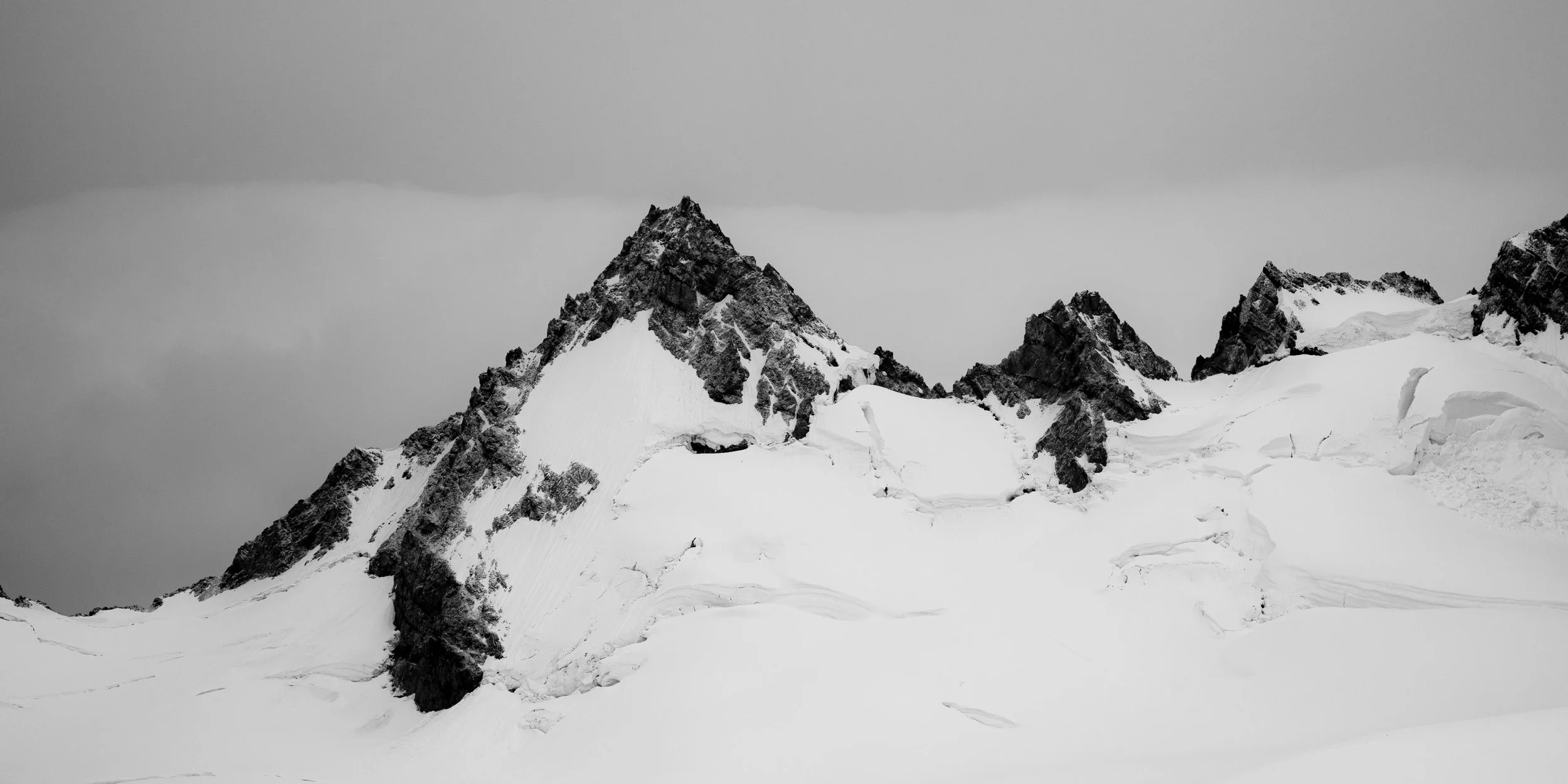 Black and white photo of snow-covered mountain peaks and slopes under a cloudy sky.