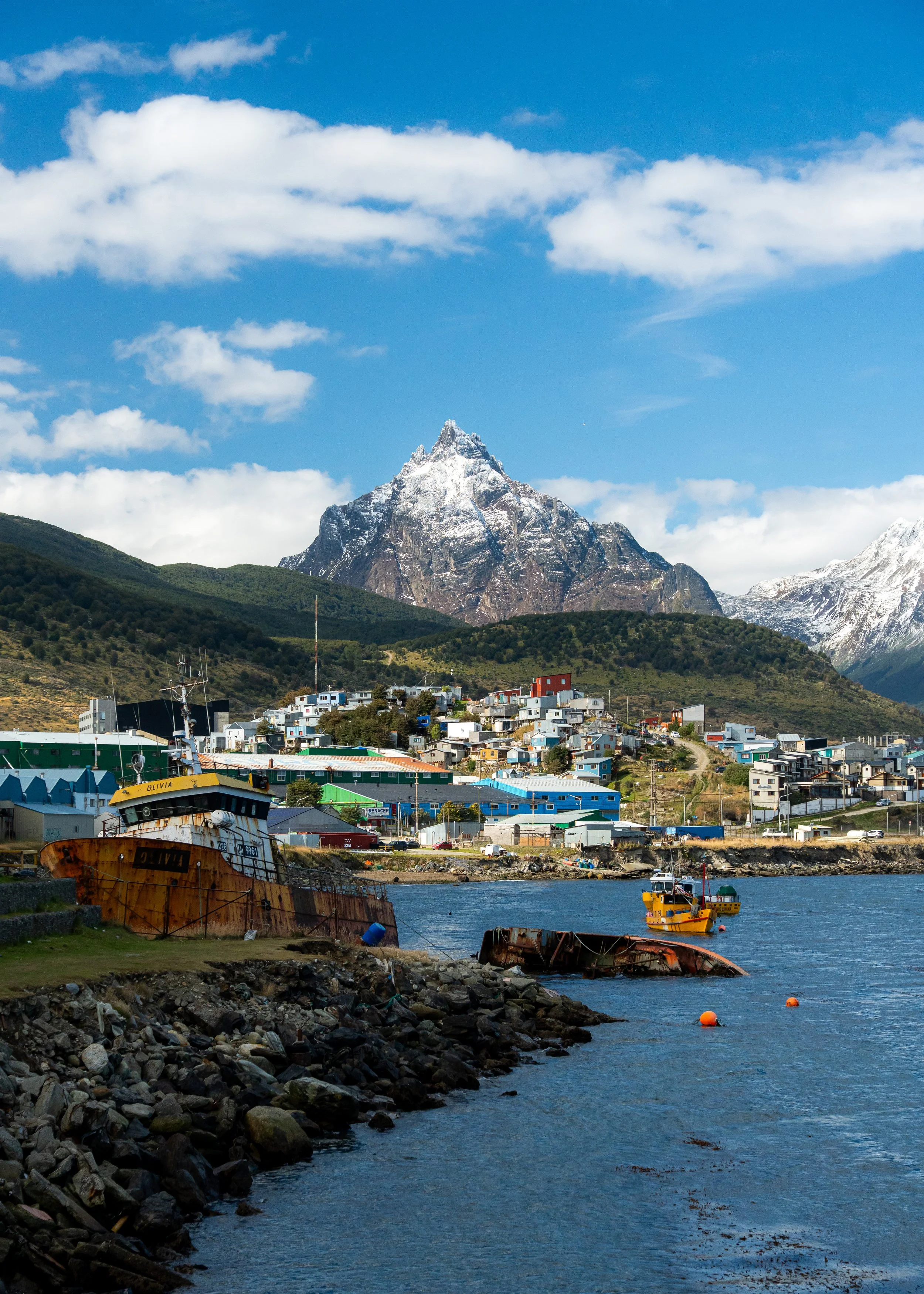 Coastal town with colorful houses at the foot of snow-capped mountains, boats docked near the shore, and a blue sky with scattered clouds.