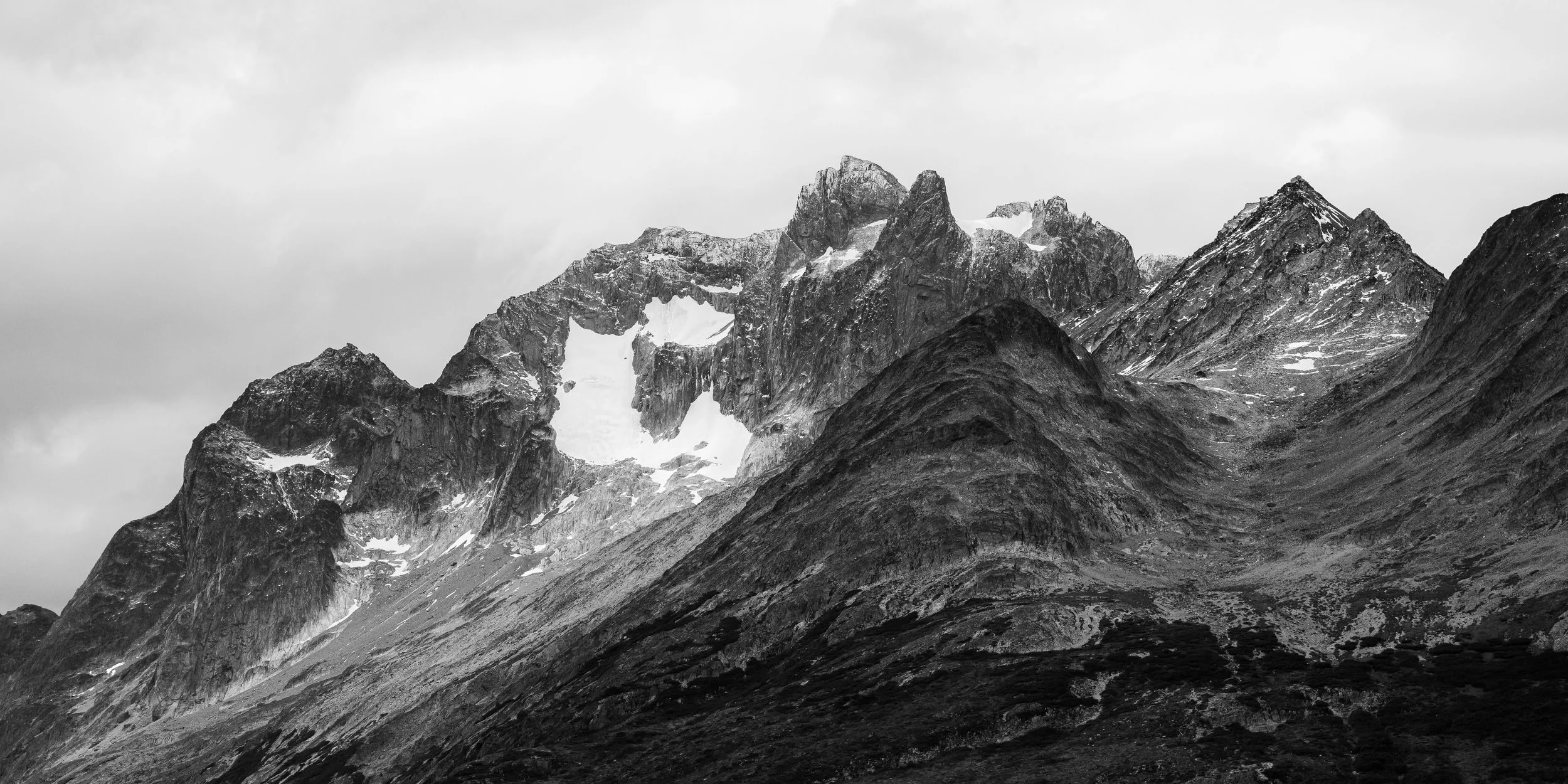Black and white photo of rugged mountain peaks with snow patches, steep slopes, and cloudy sky.